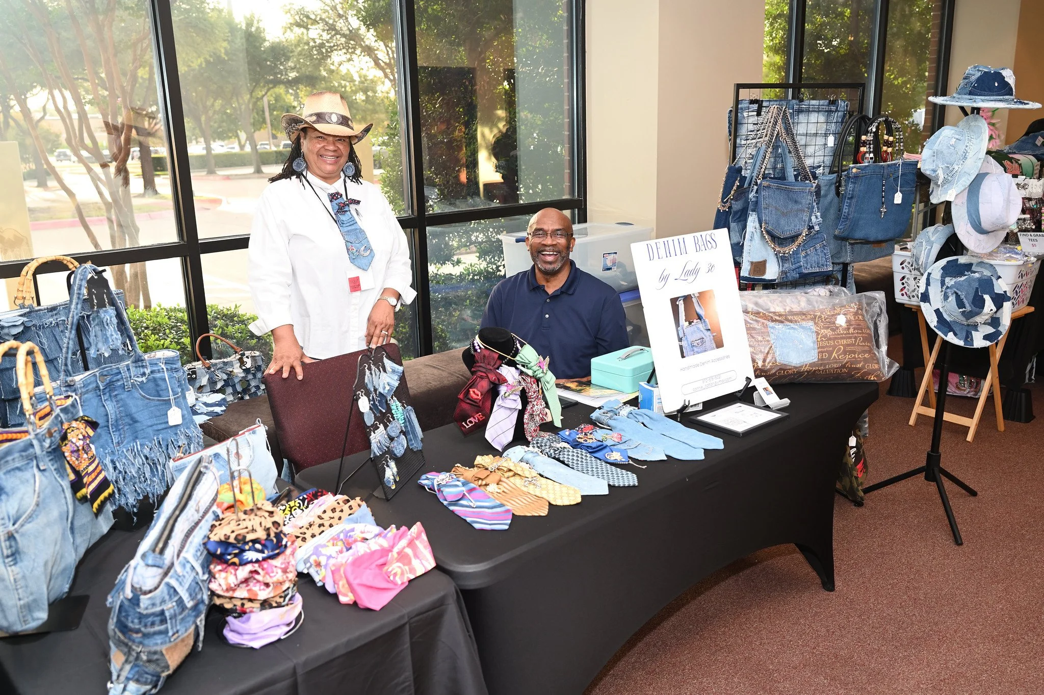 A smiling woman and man sit behind a table displaying denim and accessories at a craft fair or market. There are racks of denim bags, hats, and jewelry next to the table, with a sign that reads "Denim Bags by Lady 30." Large windows and trees are vis