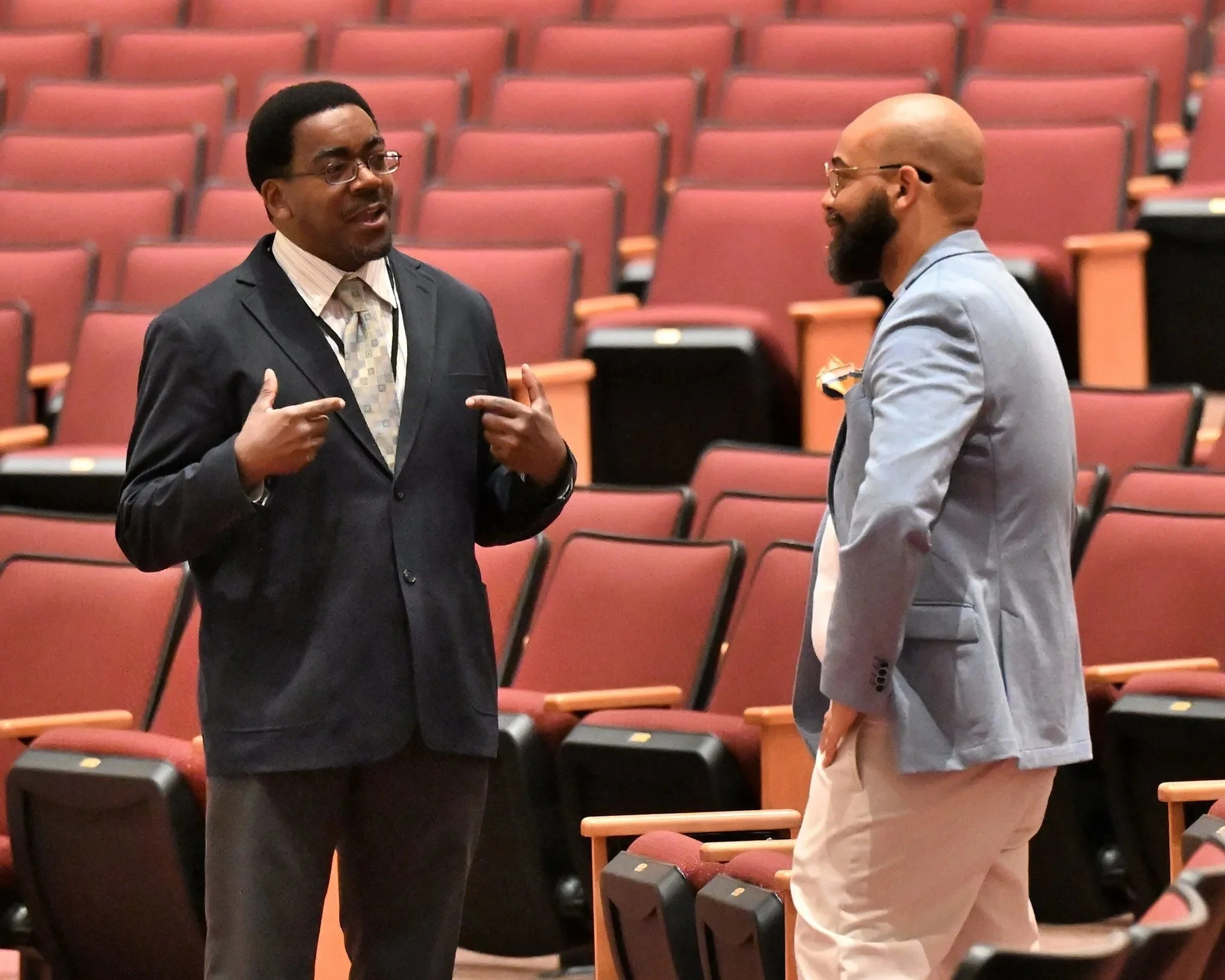 Two men engaged in conversation in an auditorium with rows of red seats. One man is dressed in a dark suit with a patterned tie, and the other is in a light gray blazer with beige pants and has a bald head and a beard.