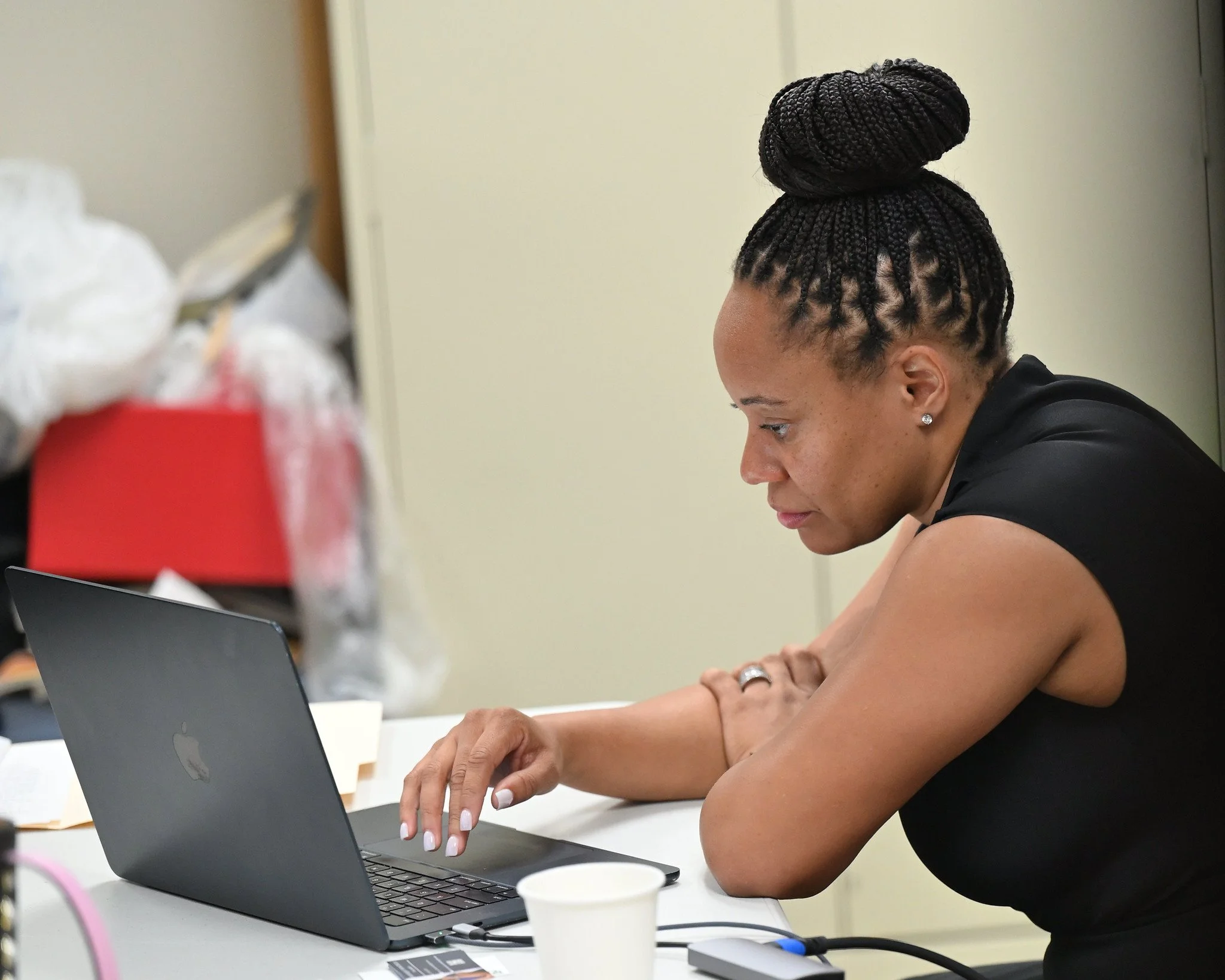 A woman with dark braided hair styled in a high bun, wearing a black sleeveless top and earrings, working on a silver MacBook laptop in a room with beige walls, surrounded by office supplies and cups.