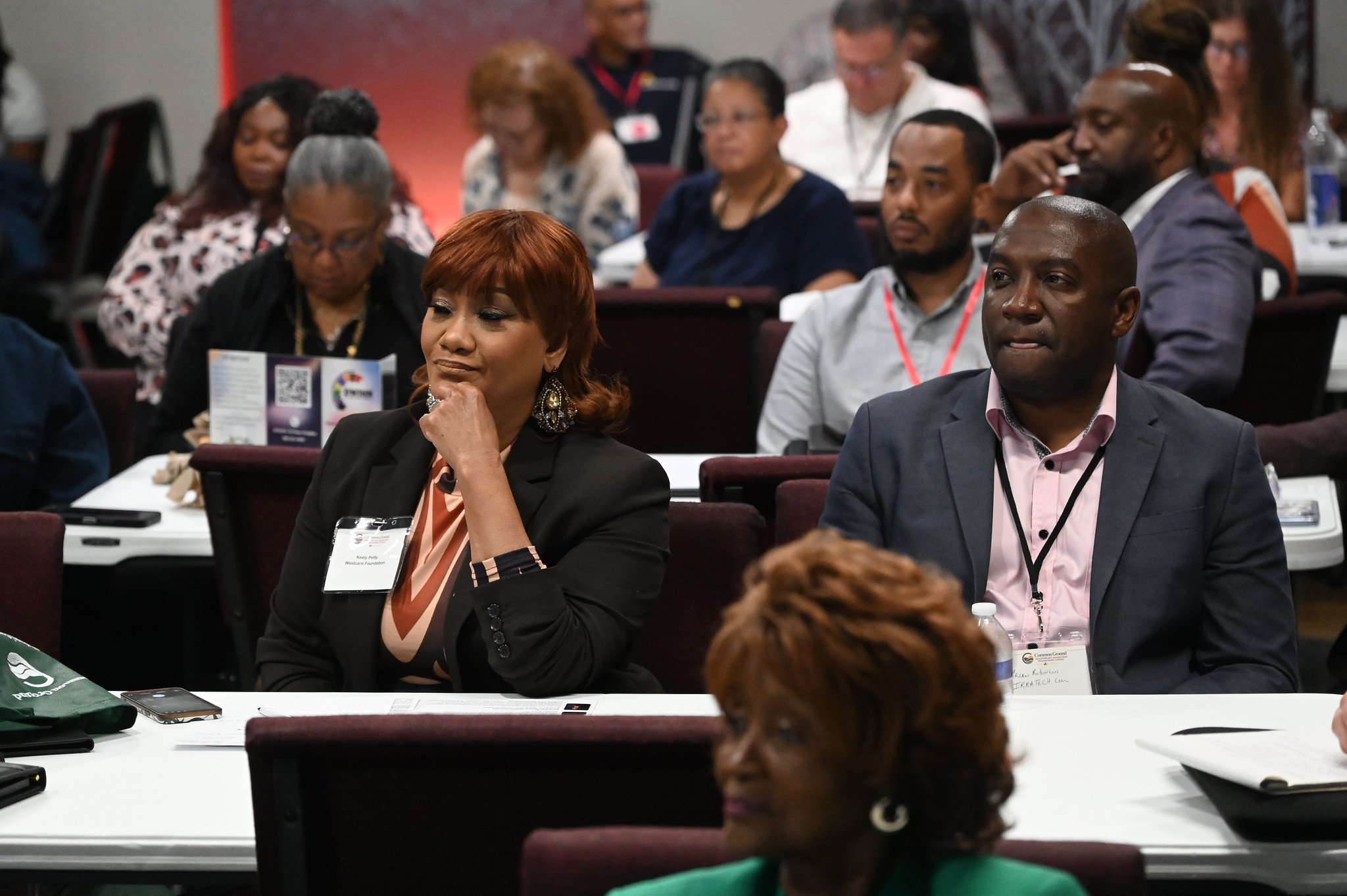 A diverse group of people attending a conference or seminar, seated at tables with conference materials, in a well-lit room.