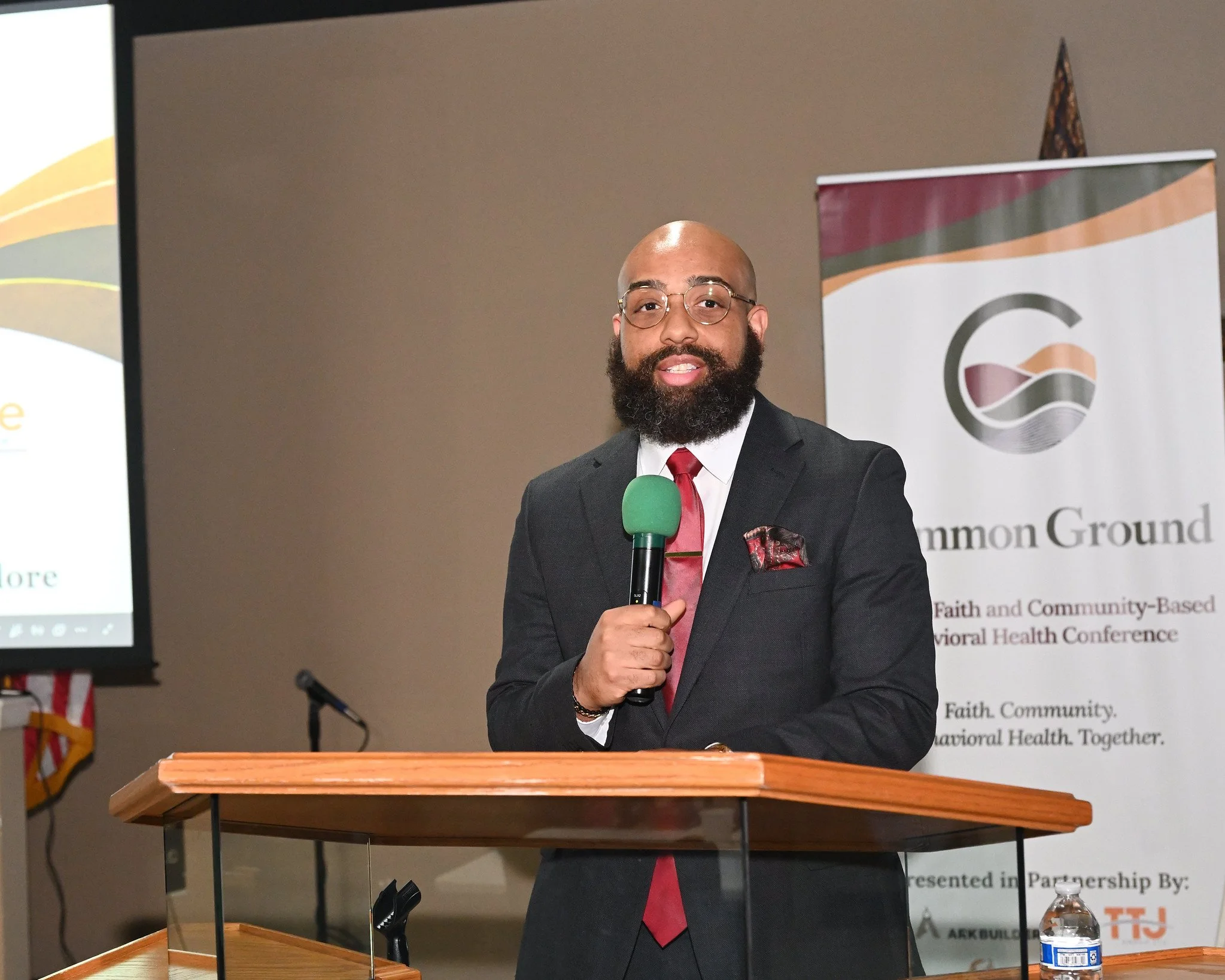 Man with a bald head, beard, glasses, wearing a black suit, white shirt, and red tie, holding a green microphone at a podium during a conference or presentation.