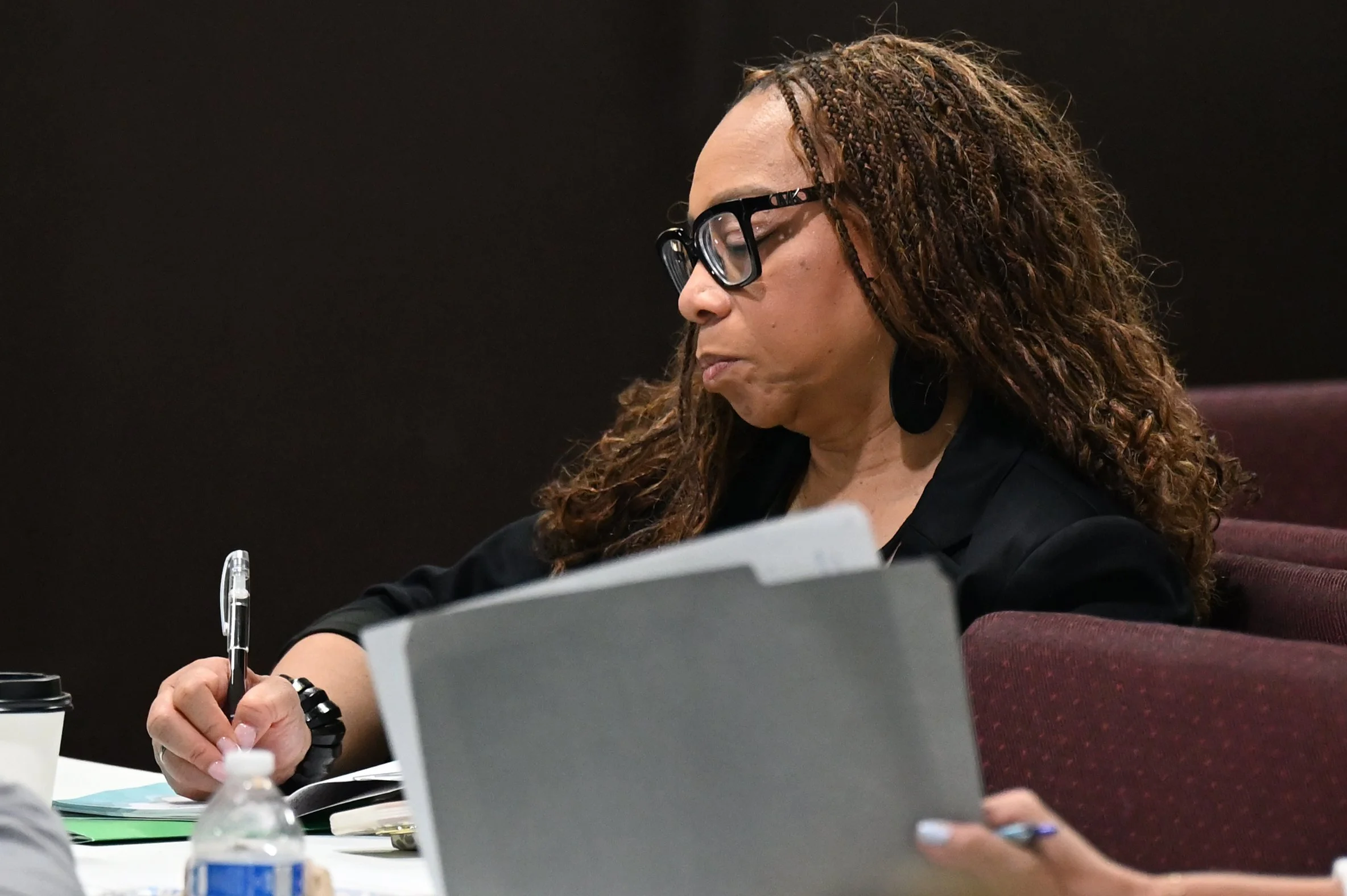 A woman with curly hair, wearing glasses, is sitting at a table, holding a pen and writing on paper, with a laptop and water bottle nearby.