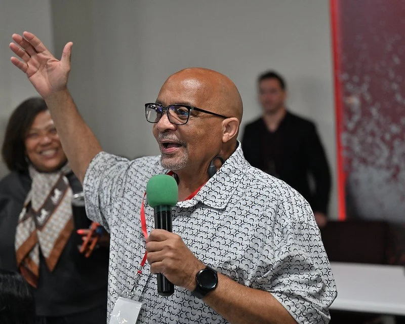 A middle-aged man with glasses, a patterned short-sleeve shirt, and a smartwatch on his left wrist, is speaking into a microphone with a green foam cover, raising his right hand, and smiling. Two women and a man are in the background, smiling in a room with a gray wall and a red banner or poster.