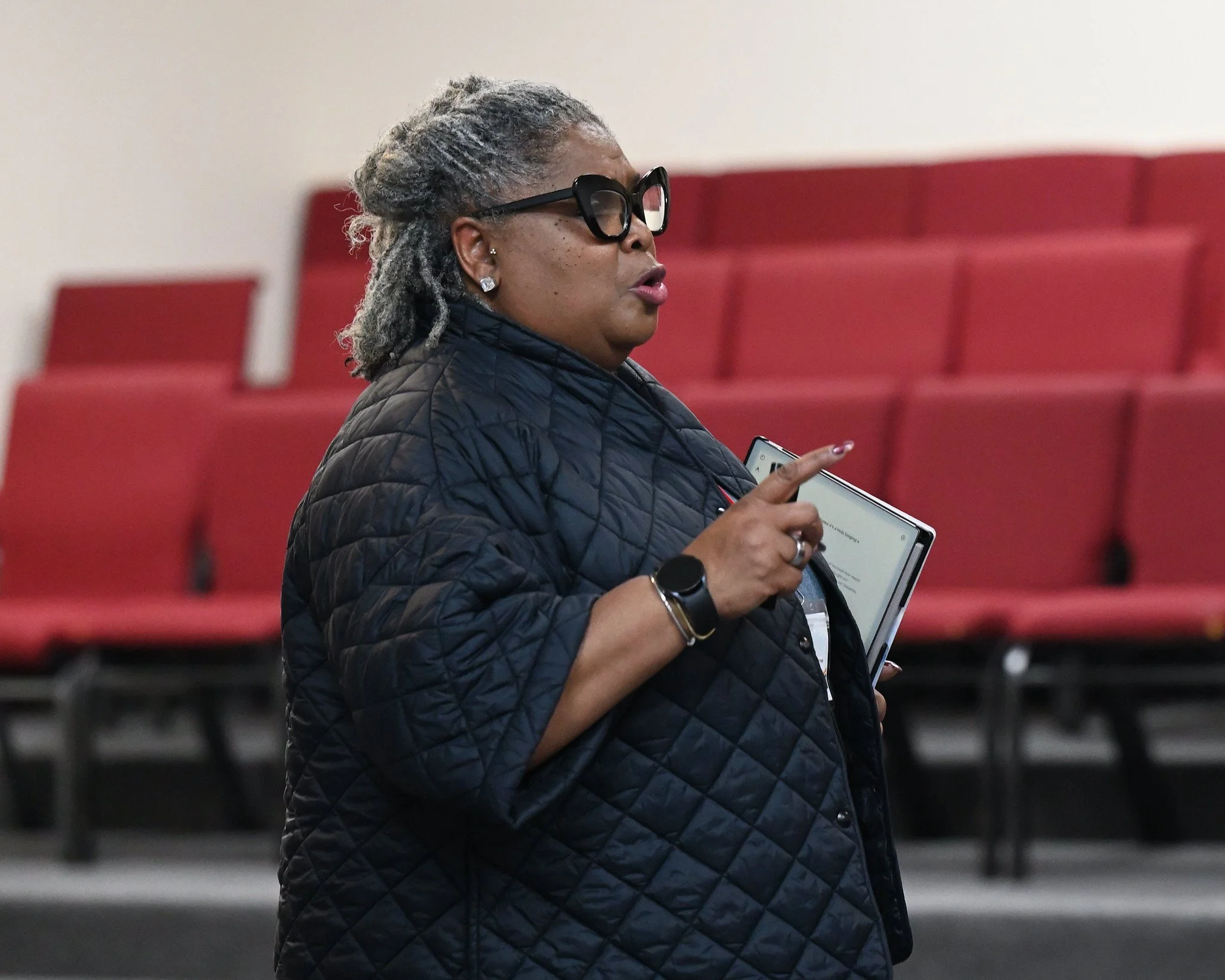 A woman with gray dreadlocks, wearing black glasses and earrings, dressed in a quilted dark jacket, is holding a smartphone and speaking in what appears to be an indoor setting with red seating.