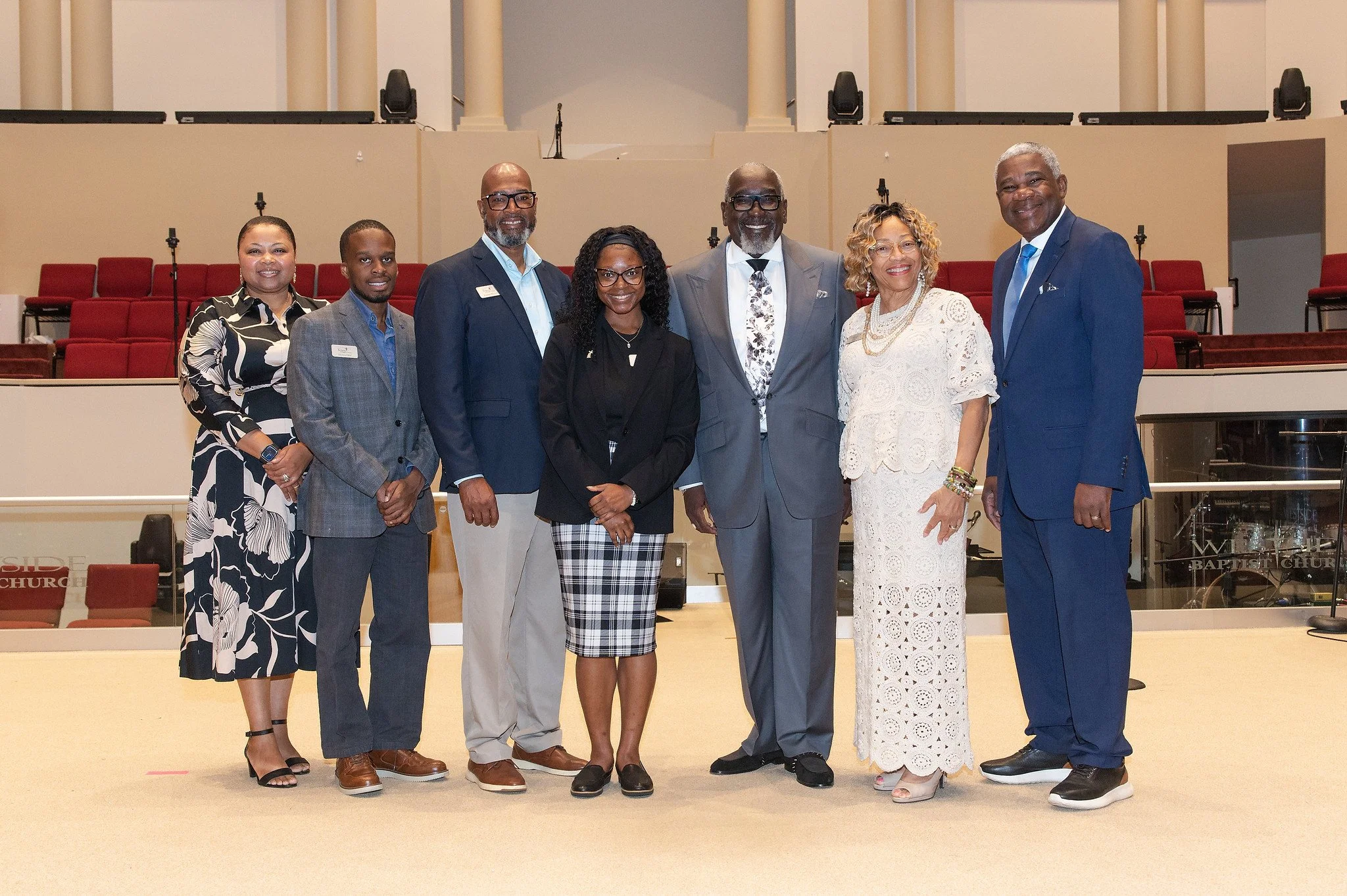 Group of seven diverse adults standing together in a church auditorium, smiling for the photo, with red chairs and audio equipment in the background.