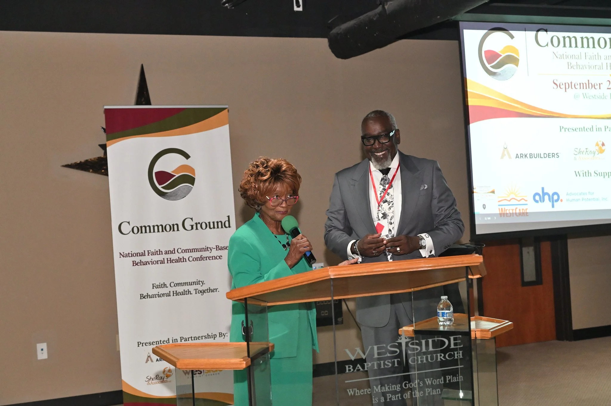 A woman in a green blazer speaking into a microphone at a podium during a conference, with a smiling man in a gray suit standing next to her. A banner with a logo and text about a faith and behavioral health conference is visible behind them.