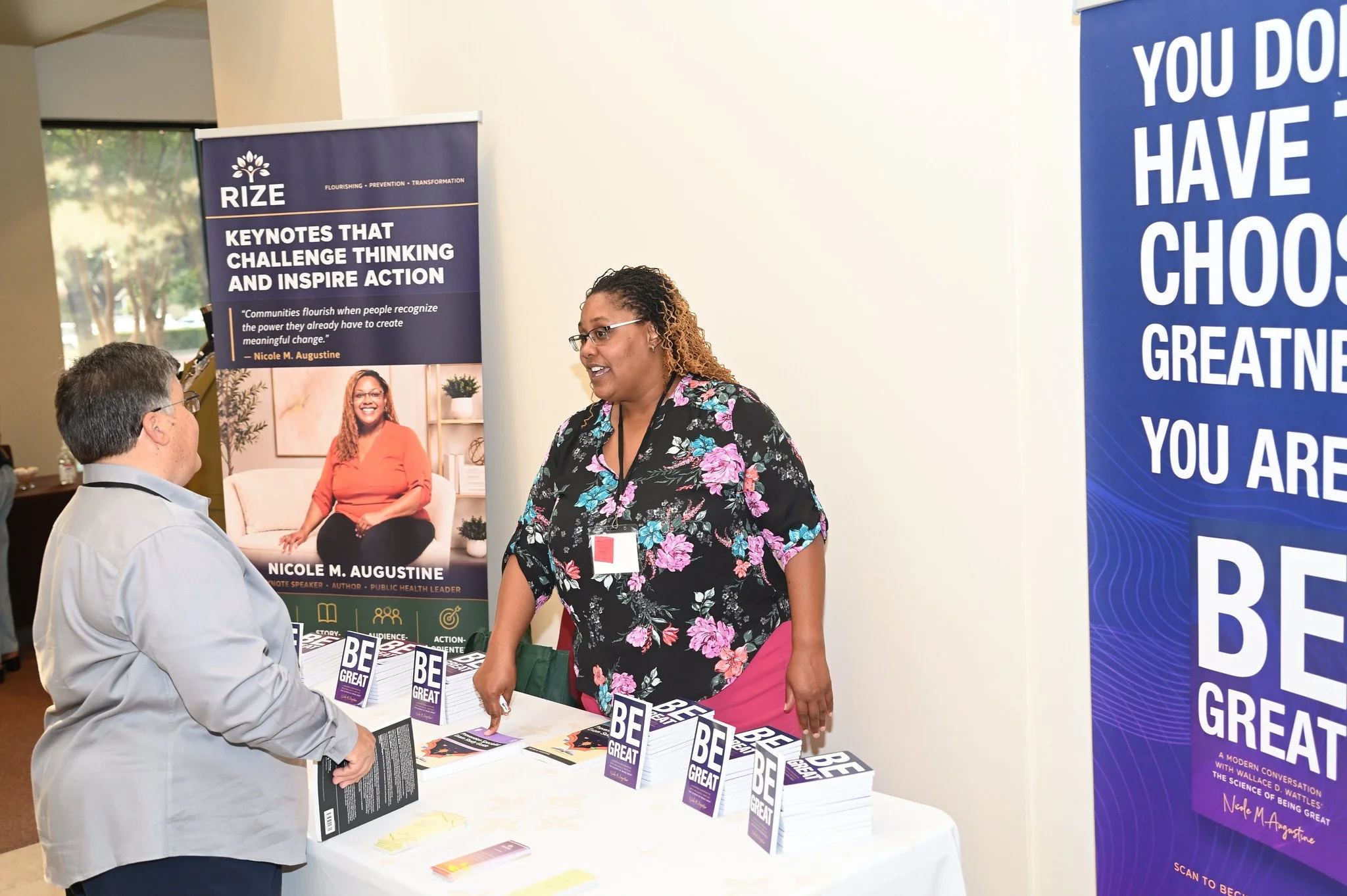A woman in a floral top and pink pants talking to a man in a light-colored shirt at a book signing event. The table displays copies of her book titled "Be Great." There are two large vertical banners behind them, one promoting the book and author Nic
