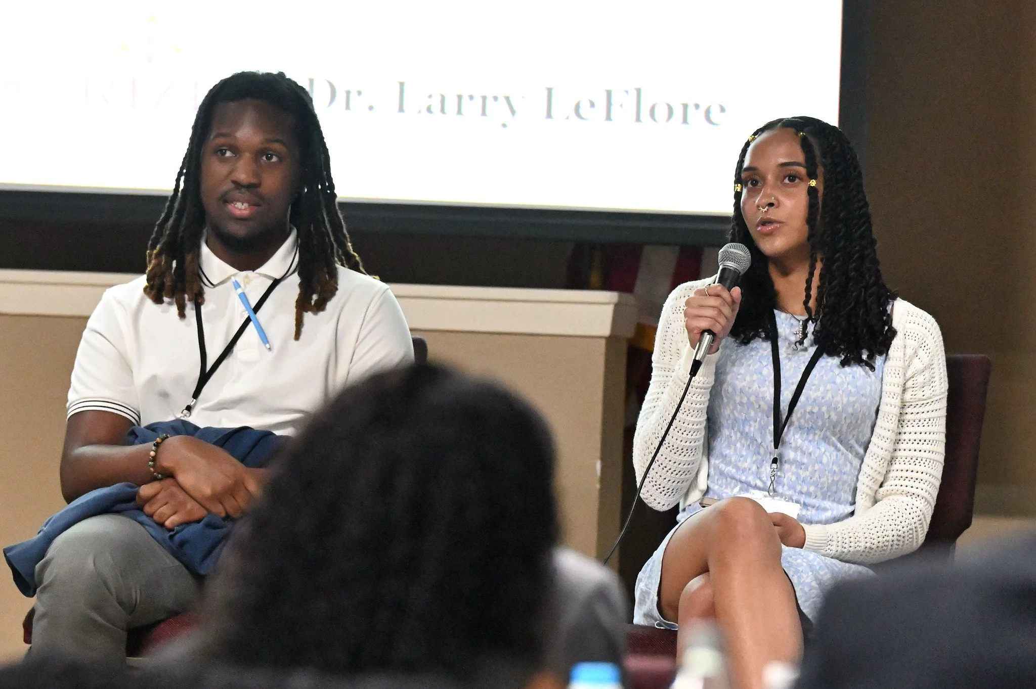Two women seated on stage at a conference, one speaking into a microphone, with a screen behind them displaying text about Dr. Larry Leflora.