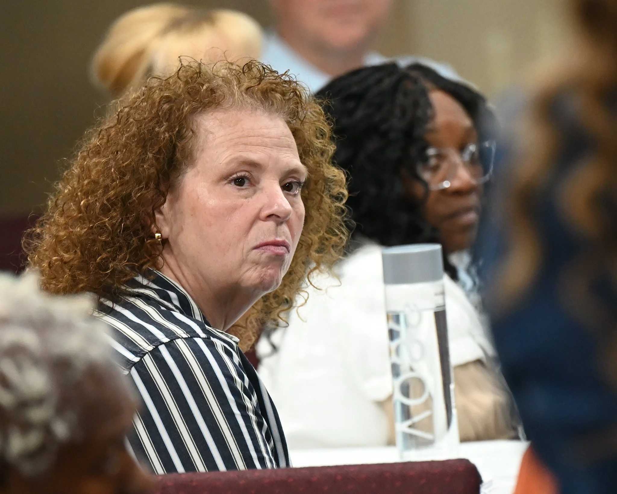 Close-up of two women sitting side by side at a meeting or conference, one with curly red hair wearing a black-and-white striped blazer, and the other with black hair and glasses, wearing a white shirt with a name badge.