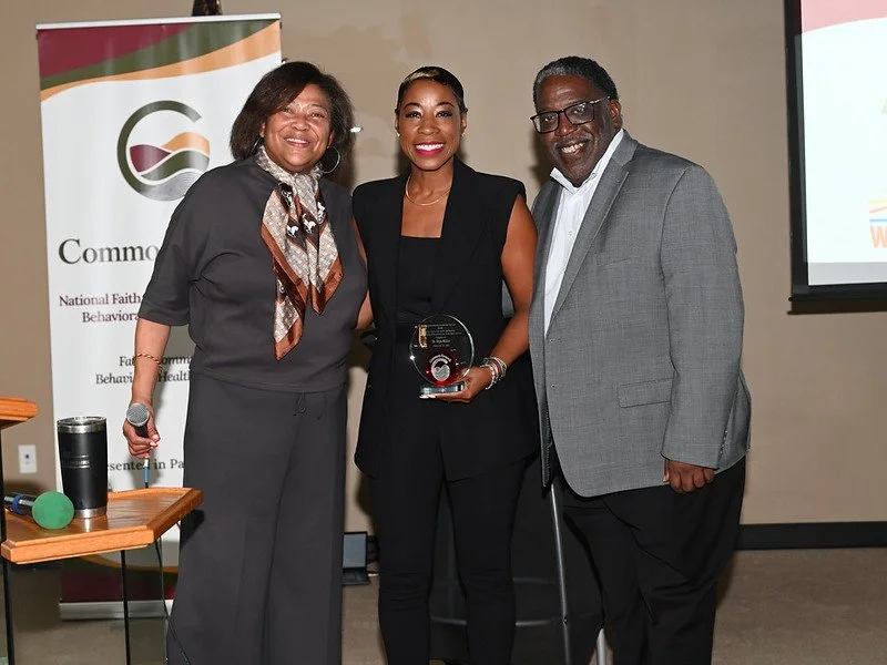 Three people standing together, smiling, with a woman in the center holding an award. There is a banner in the background and a table with a green microphone and a tumbler on it.