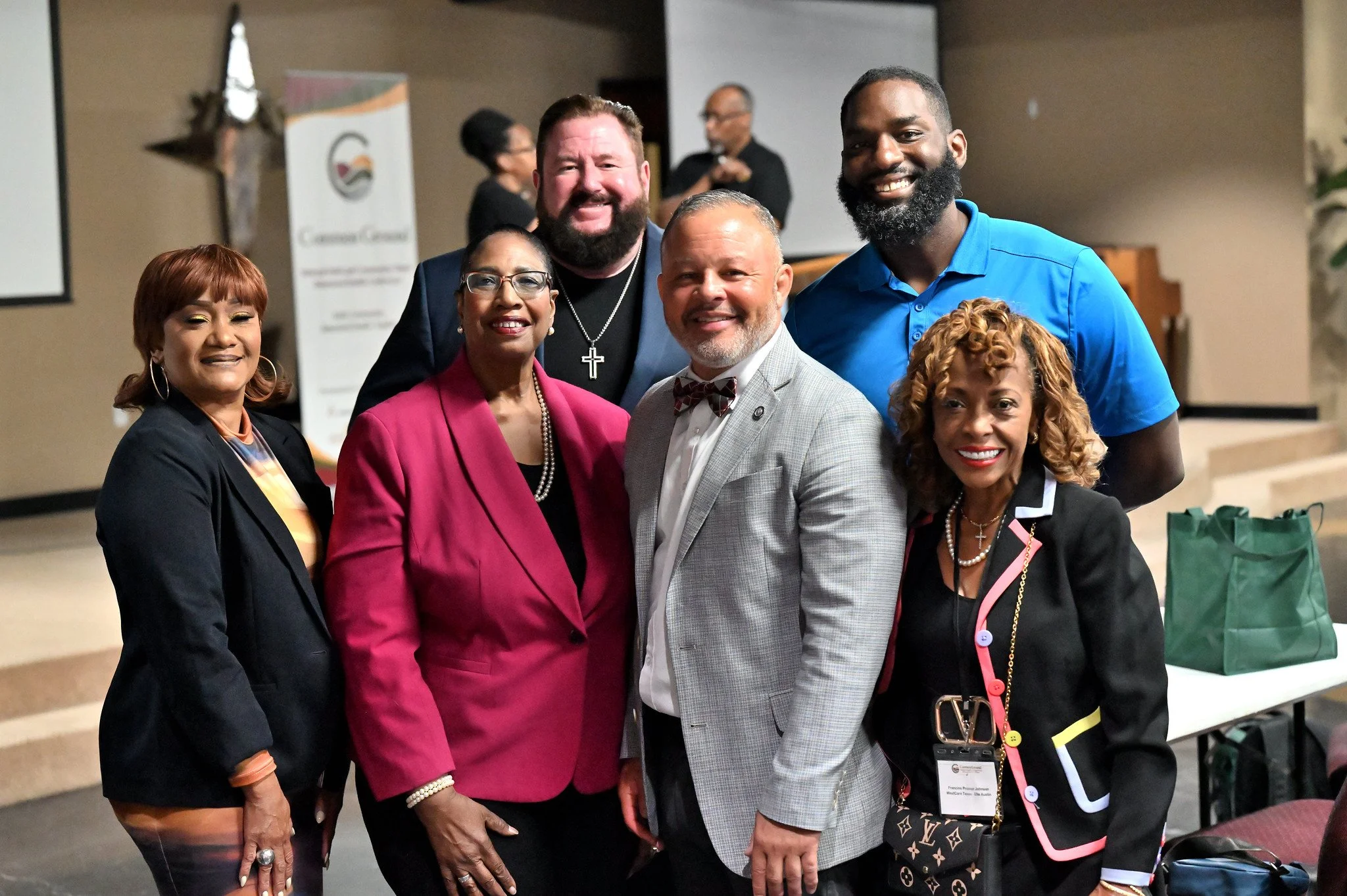A group of six diverse individuals in business attire smiling and posing together indoors at a conference or event.
