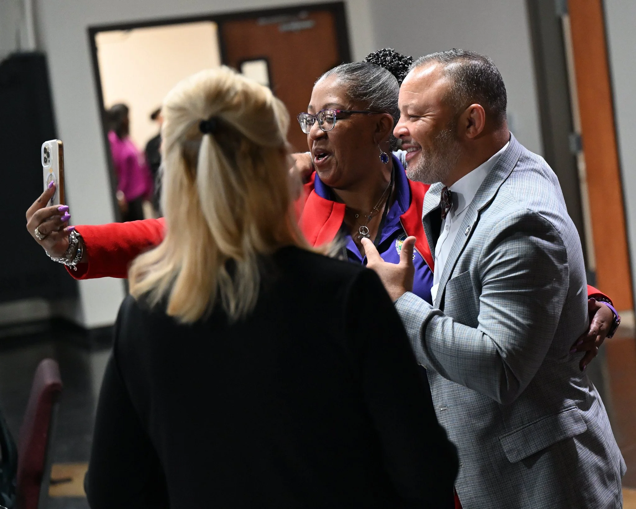Two women and a man are smiling and taking a selfie together at an indoor event. The woman on the left has blonde hair in a ponytail, and the woman in the middle has dark hair and is wearing glasses and earrings. The man on the right has short, gray 