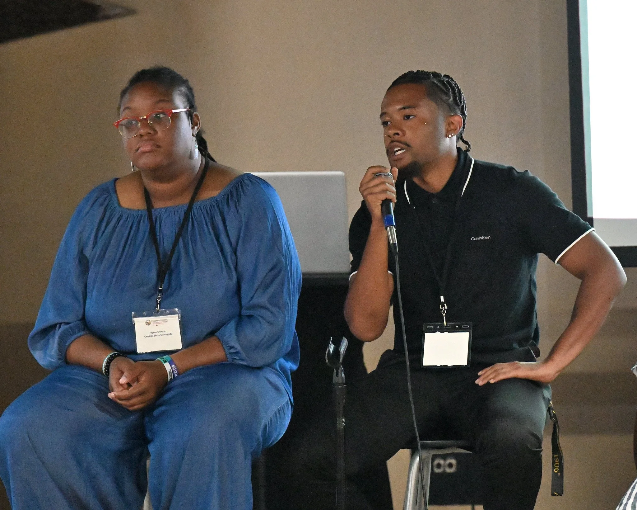 Two women are seated at a conference, one with glasses and a blue blouse with a name badge, the other with braided hair, speaking into a microphone, with a presentation screen in the background.