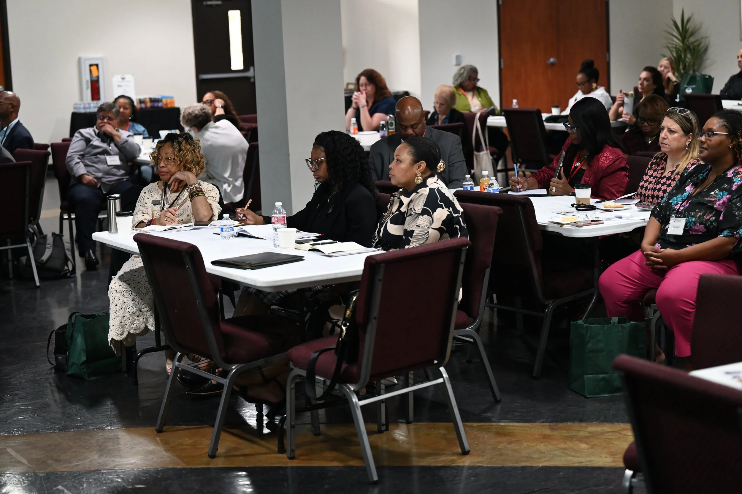 A group of people attending a conference or seminar sitting at round tables with notebooks, drinks, and pens, in a room with beige walls and dark flooring.