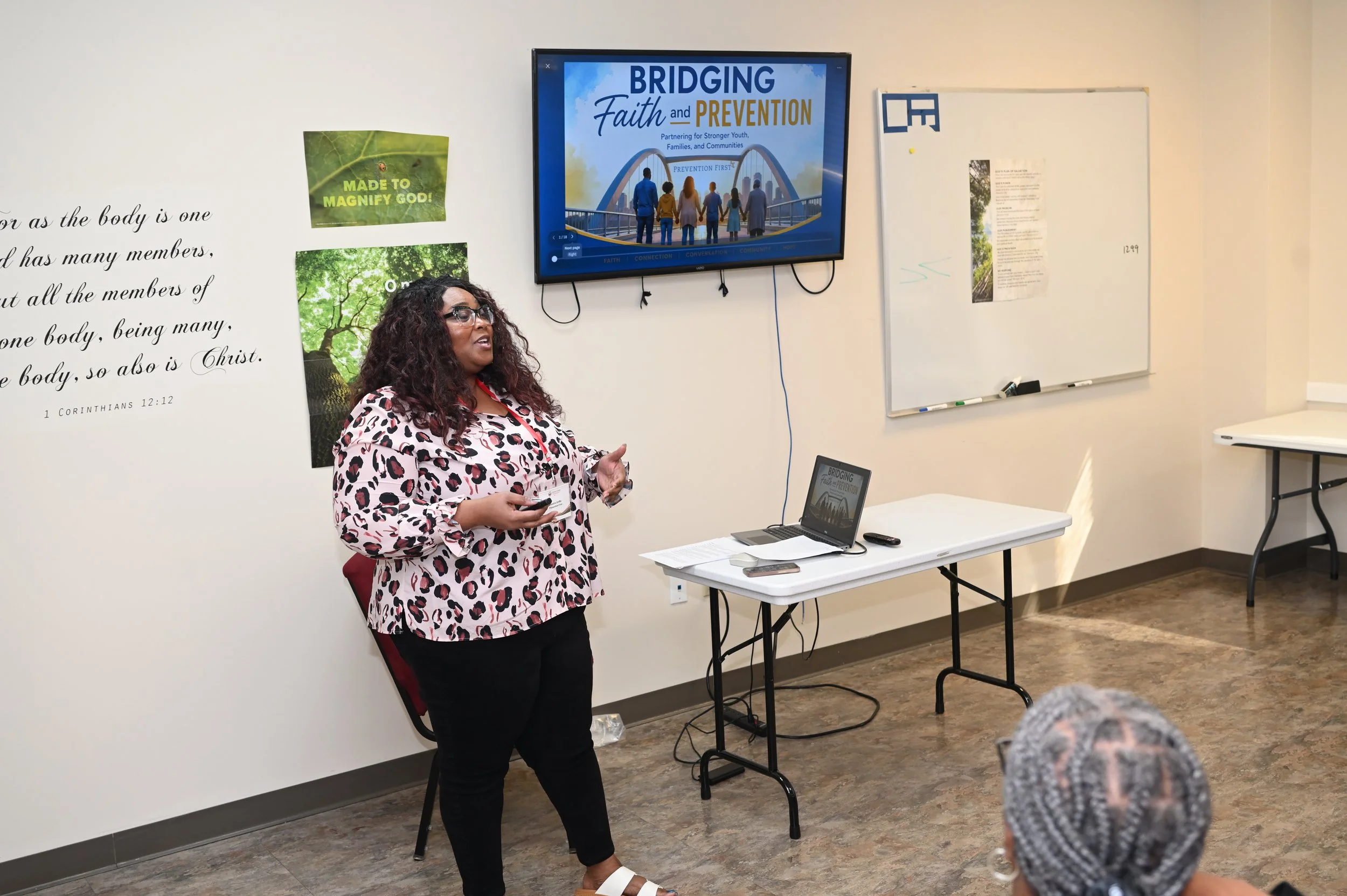 A woman with curly dark hair, glasses, and a leopard print blouse is standing and speaking in front of a classroom. Behind her is a whiteboard and a large screen displaying a presentation titled 'Bridging Faith and Prevention.' Several posters and writings are on the classroom wall, and there are students listening, some with gray hair.