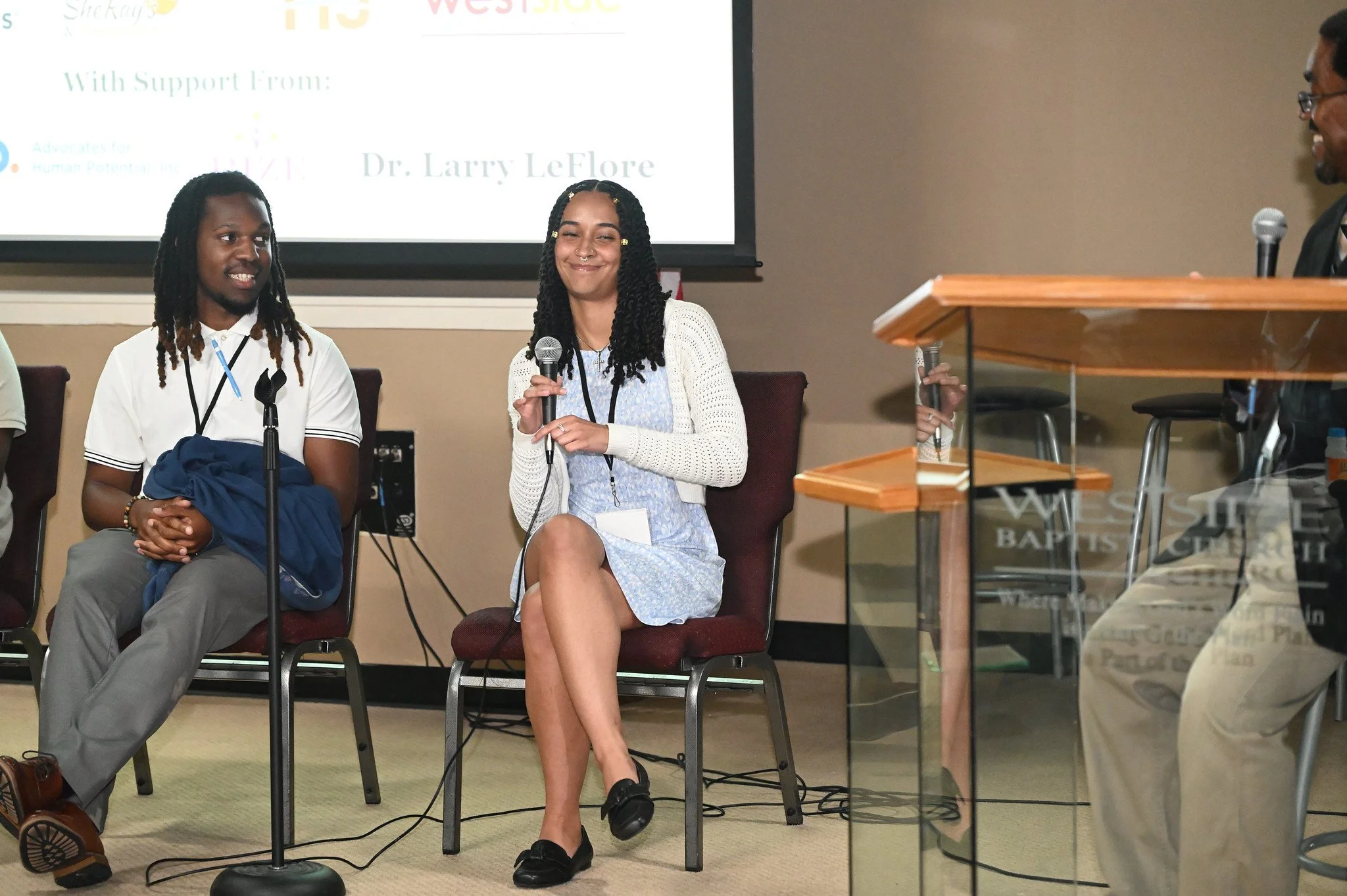 Two women sitting on chairs during a panel discussion or presentation, with a man at a podium on the right. One woman is holding a microphone and smiling, wearing a light blue dress and a white sweater, while the other woman has long dreadlocks, is h