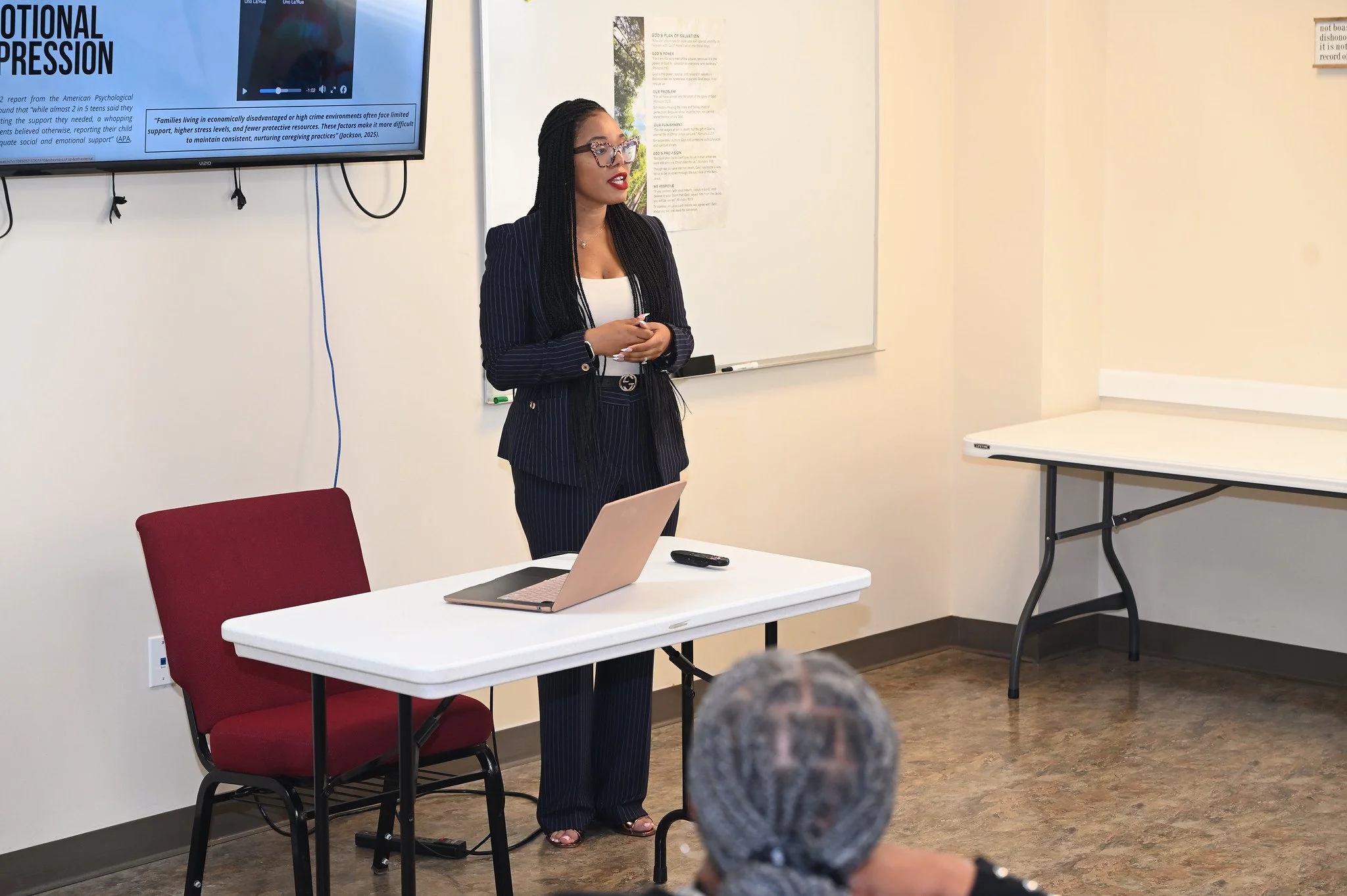 A woman in a pinstripe suit with glasses and long braided hair giving a presentation in a classroom or conference room. She is standing next to a whiteboard and a TV screen displaying text and a video. A laptop and remote are on the table in front of