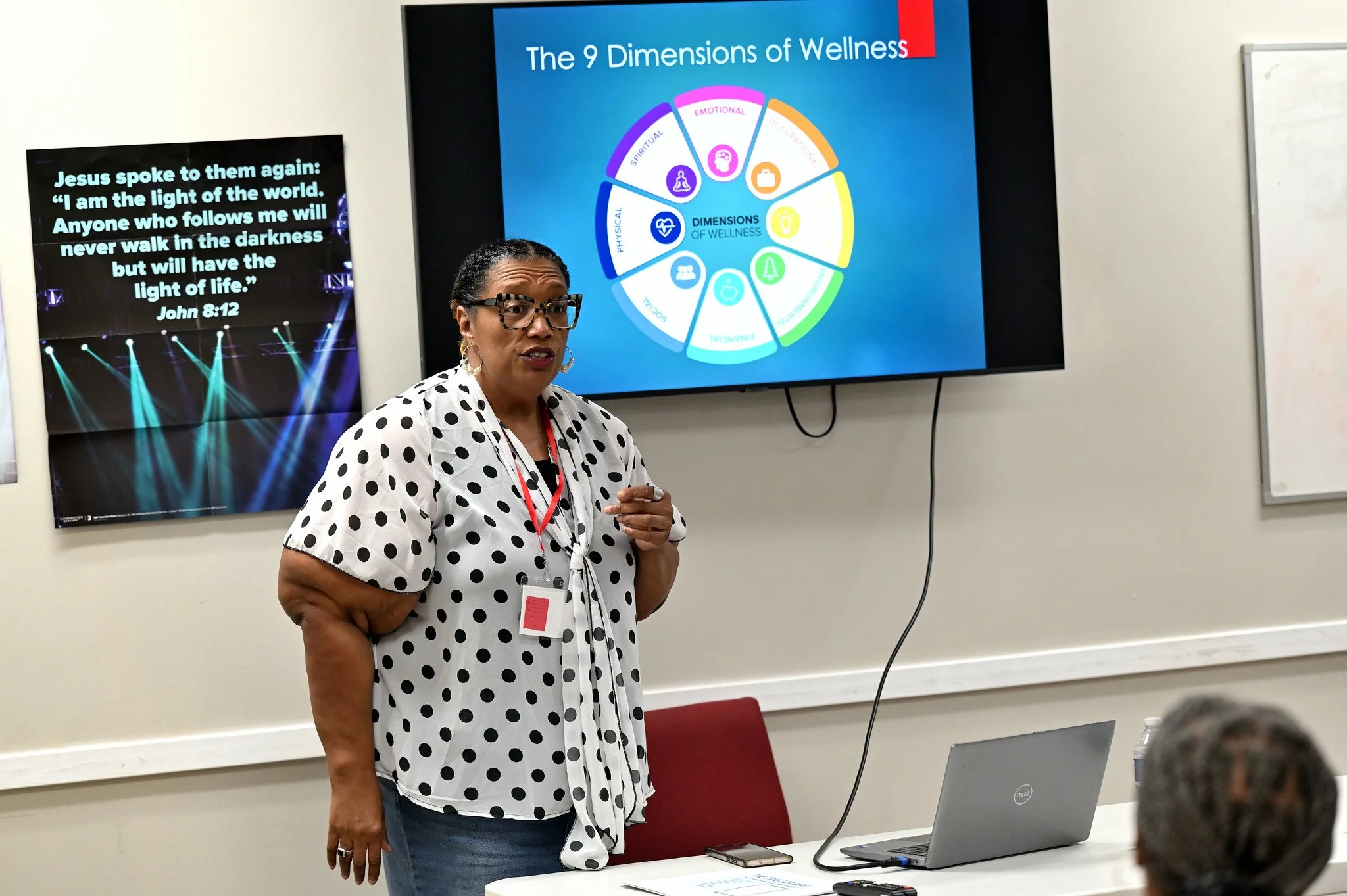 A woman with black glasses and a polka dot shirt giving a presentation in a classroom or conference room. Behind her, on a large monitor, there is a slide titled 'The 9 Dimensions of Wellness' with a colorful wheel diagram. To her left, another scree