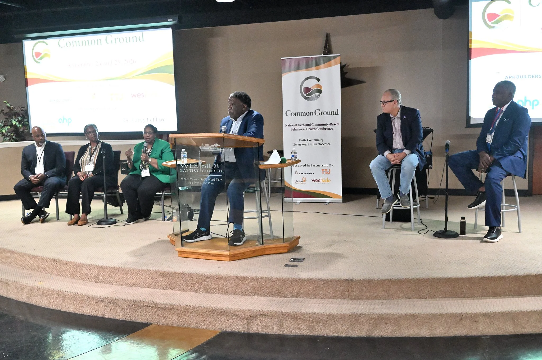 A panel of six diverse individuals seated and standing on a stage at a conference, with two large screens displaying the event title 'Common Ground' and a banner with the same logo and details in the background.