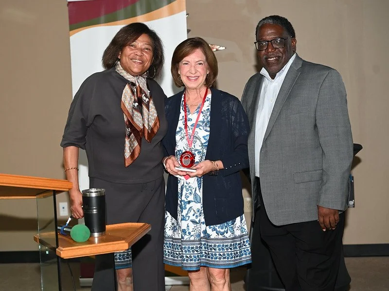 Three women and one man standing together at an award ceremony, with one woman holding a glass award. The woman on the left is wearing a dark outfit with a patterned scarf, the woman in the middle is in a blue floral dress with a medal around her nec