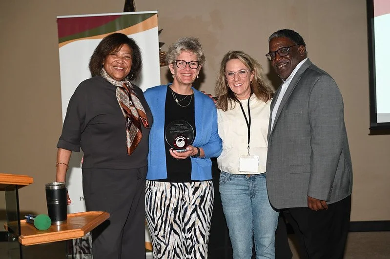 Four people standing together at an awards ceremony, three women and one man, smiling. The woman second from the left holds a plaque or award. They are indoors, with a screen and a banner in the background.