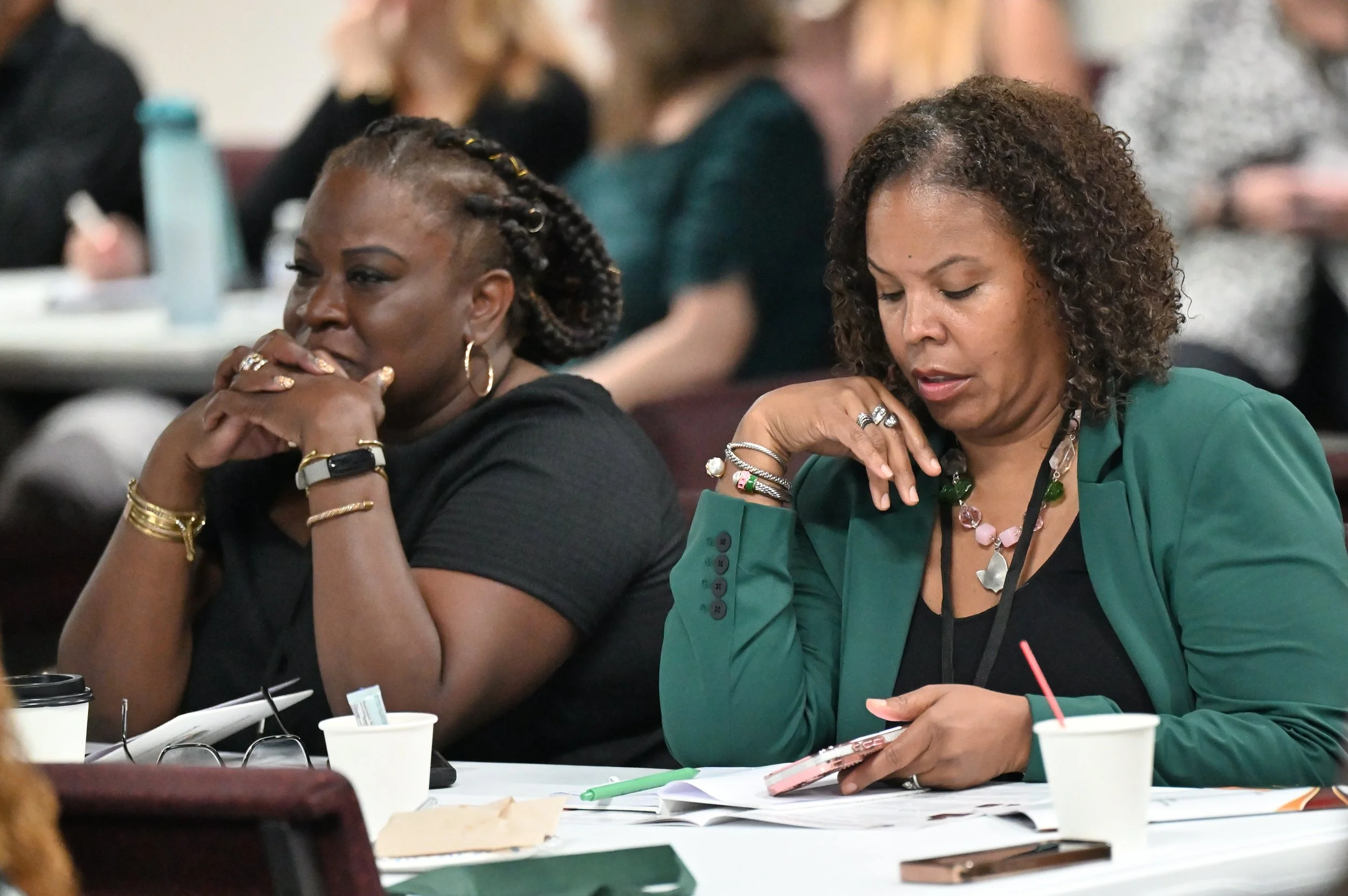 Two women seated at a table, looking down with thoughtful expressions, surrounded by papers, notebooks, cups, and pens, in a conference or meeting setting.