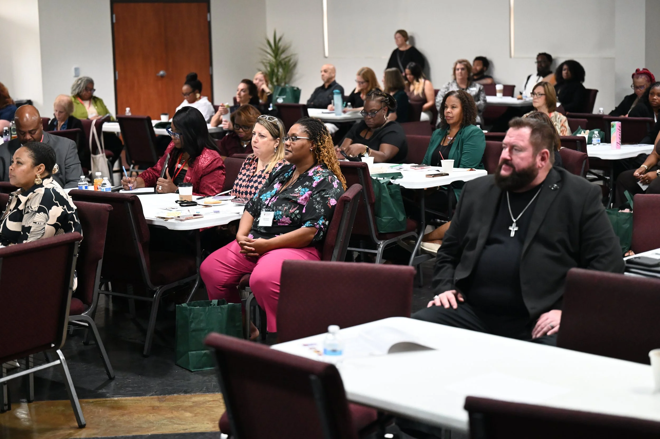 A diverse group of people seated at tables in a conference room, attentively listening to a speaker or presentation, with some taking notes and others holding cups.