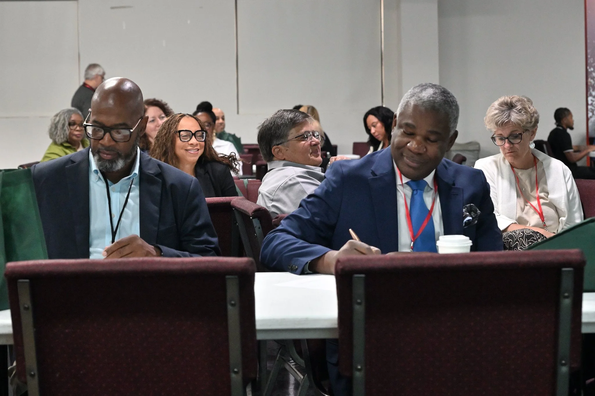 A group of diverse individuals seated in a conference room, engaging in a professional event or seminar, with some taking notes and others listening attentively.