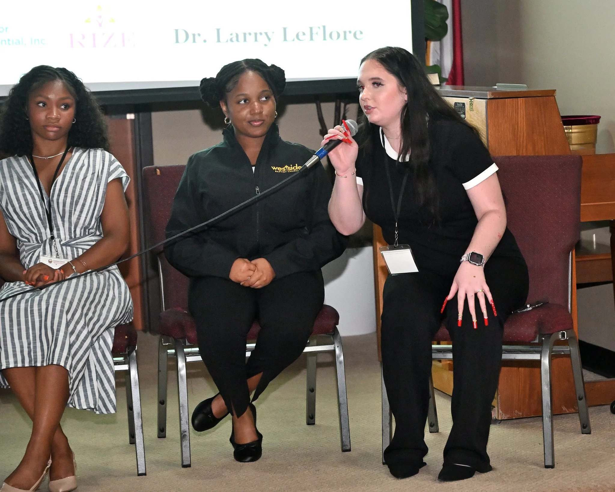 Three women seated on chairs. The woman on the right is speaking into a microphone, holding it with her right hand. The woman in the middle is wearing a black jacket with a logo that reads 'Westside Baptist Church' and has her hands clasped in her la