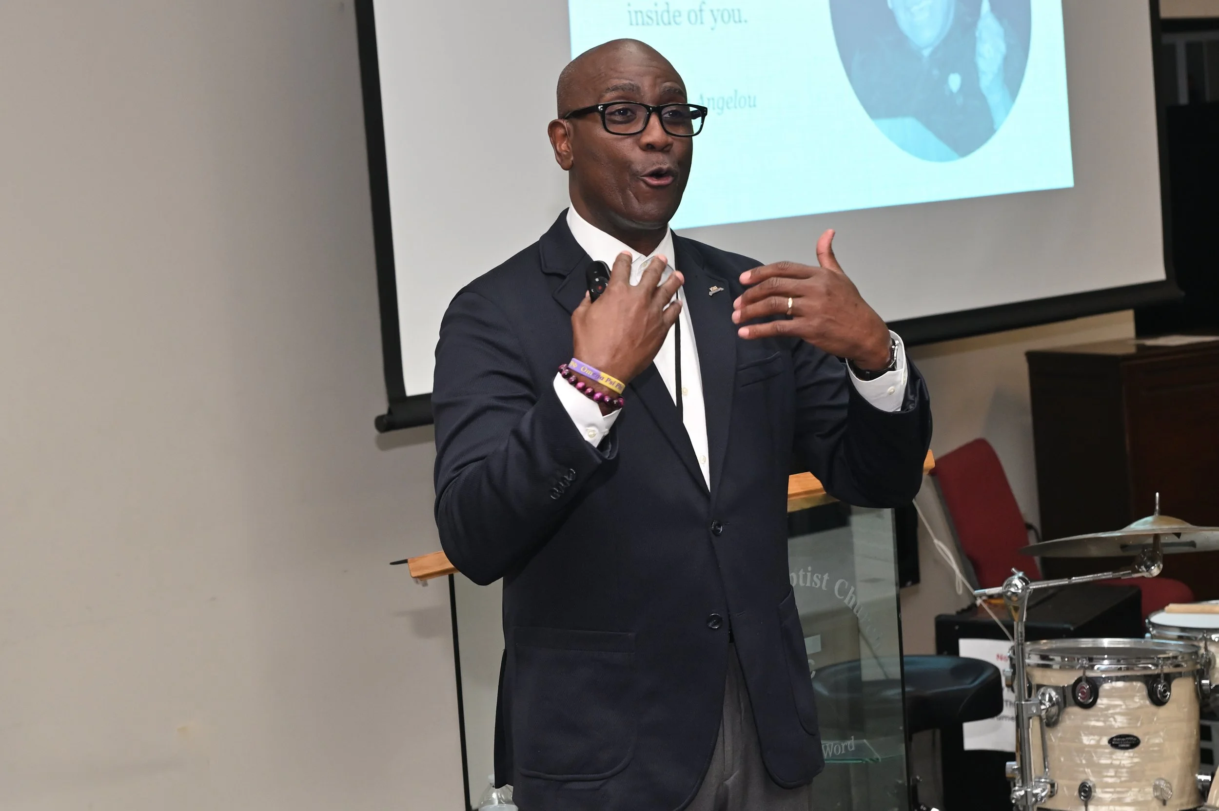 A man in a dark suit and glasses appears to be giving a presentation, standing in front of a projector screen with text and an image. He is speaking and gesturing with his hands.
