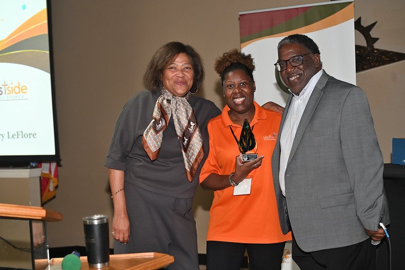 Three people standing together at an indoor event. The woman in the middle, wearing an orange shirt, is holding a glass award. The other woman and man are smiling, dressed in business attire, with a presentation screen in the background.