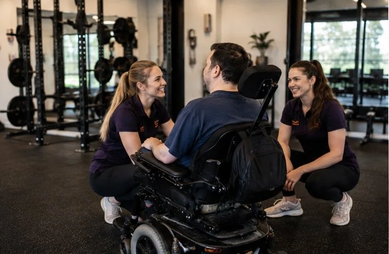 Two women and a man in a wheelchair smiling and talking in a gym, with gym equipment in the background.