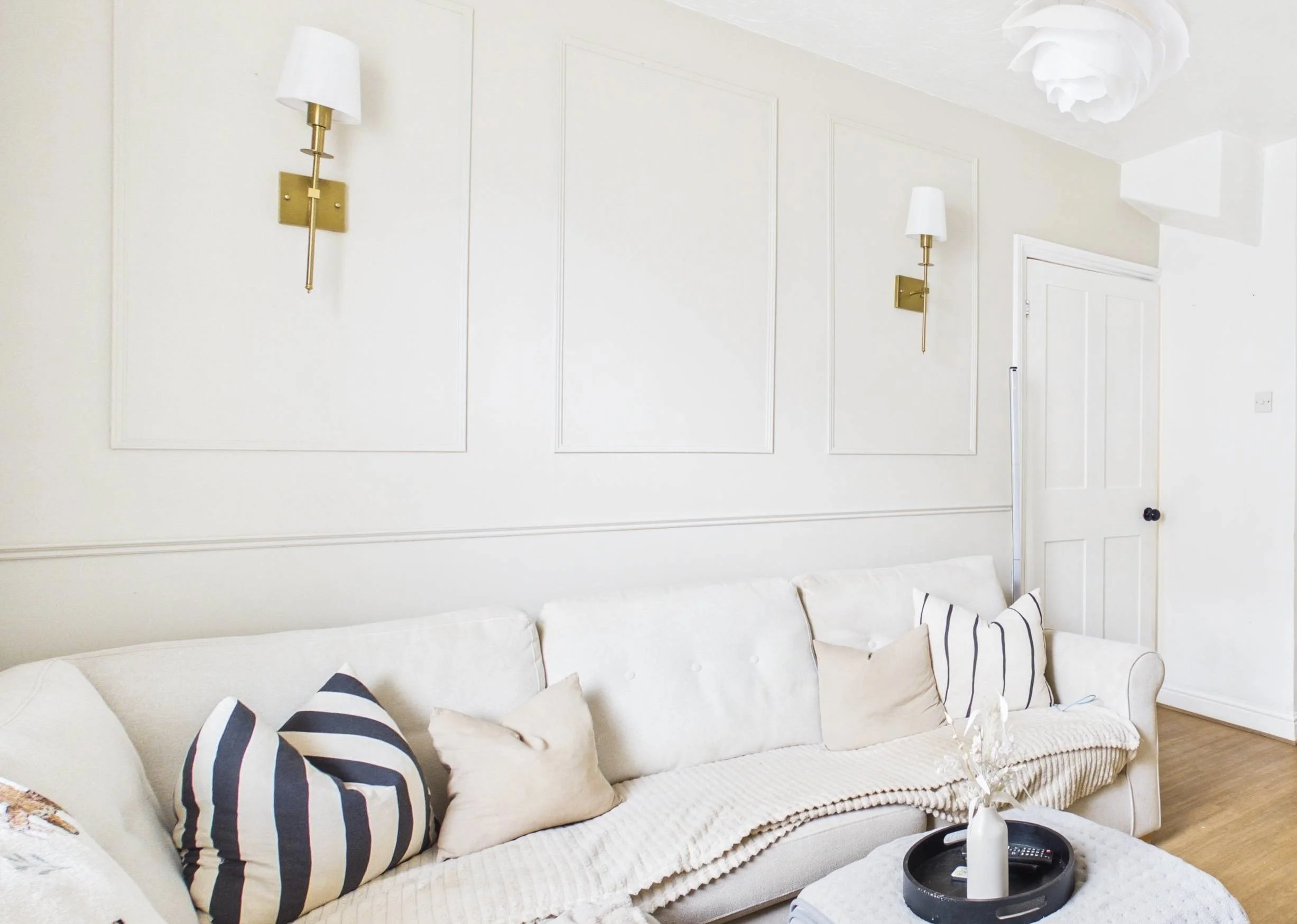 Living room with a white sofa decorated with striped and solid-colored cushions, and a black tray with a white candle and remote control on a round table in front.