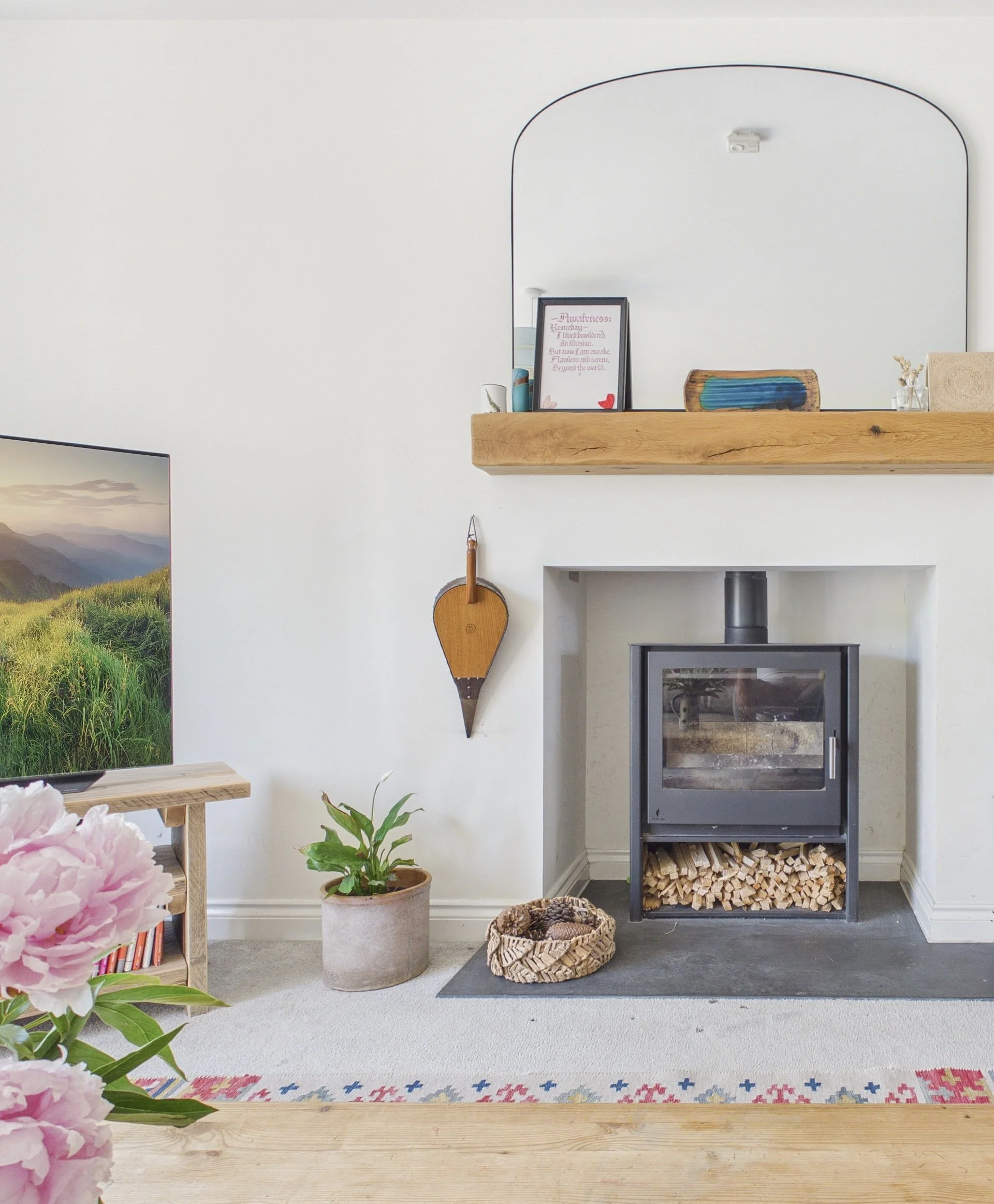 Living room with white walls, a wooden shelf, a small fireplace, a plant in a beige pot, a basket of firewood, a colorful woven basket, and pink flowers in the foreground.