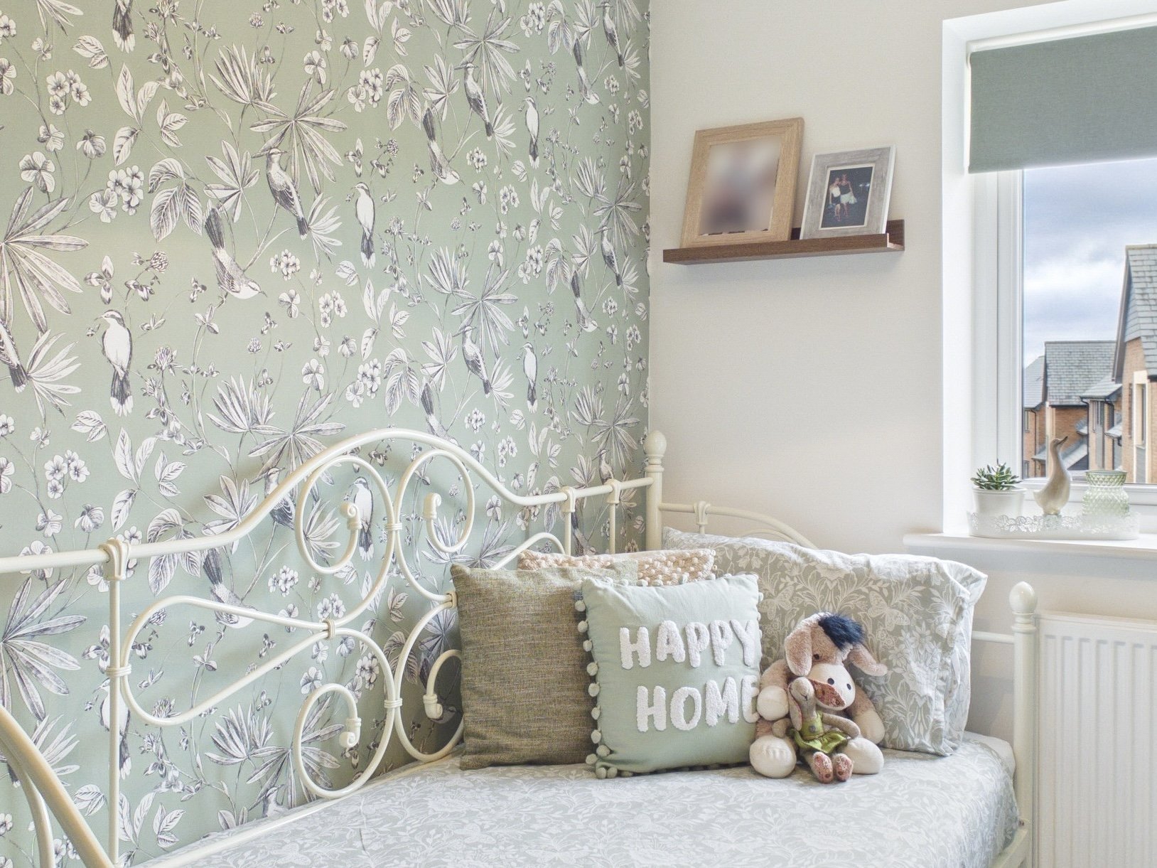 A cozy bedroom corner featuring a white, vintage-style metal bed with decorative pillows, including one with "HAPPY HOME" written on it, and plush stuffed animals. The wall behind the bed has a green botanical and bird patterned wallpaper, and a white window sill displays potted plants and decorative items, with a view of rooftops outside.