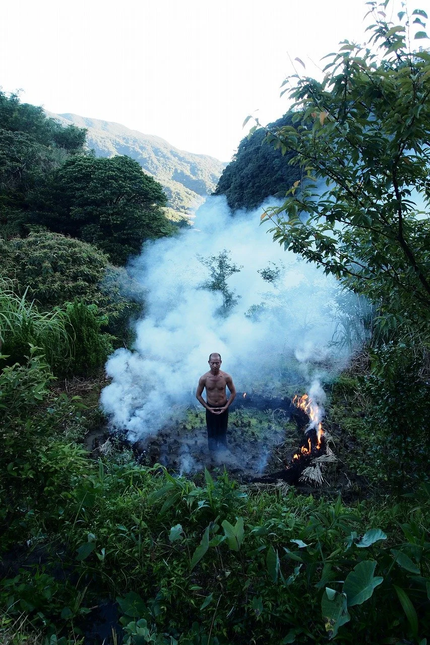 A shirtless man standing in a smoky, lush green forest near a small fire, with mountains in the background.