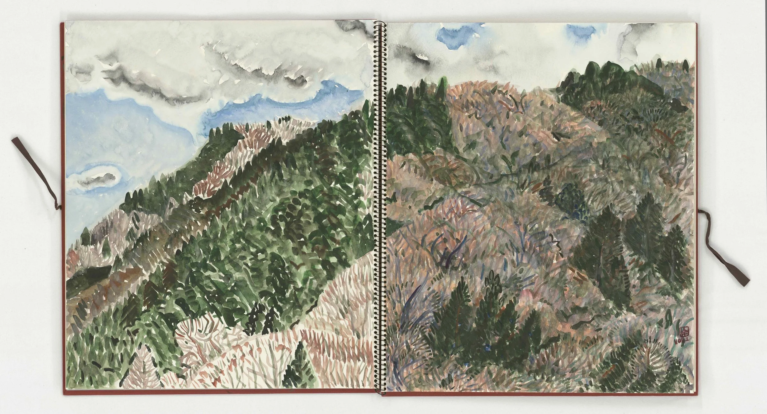 An open sketchbook with a watercolor painting of a mountain landscape, featuring cloudy sky, green trees, and pinkish hills.