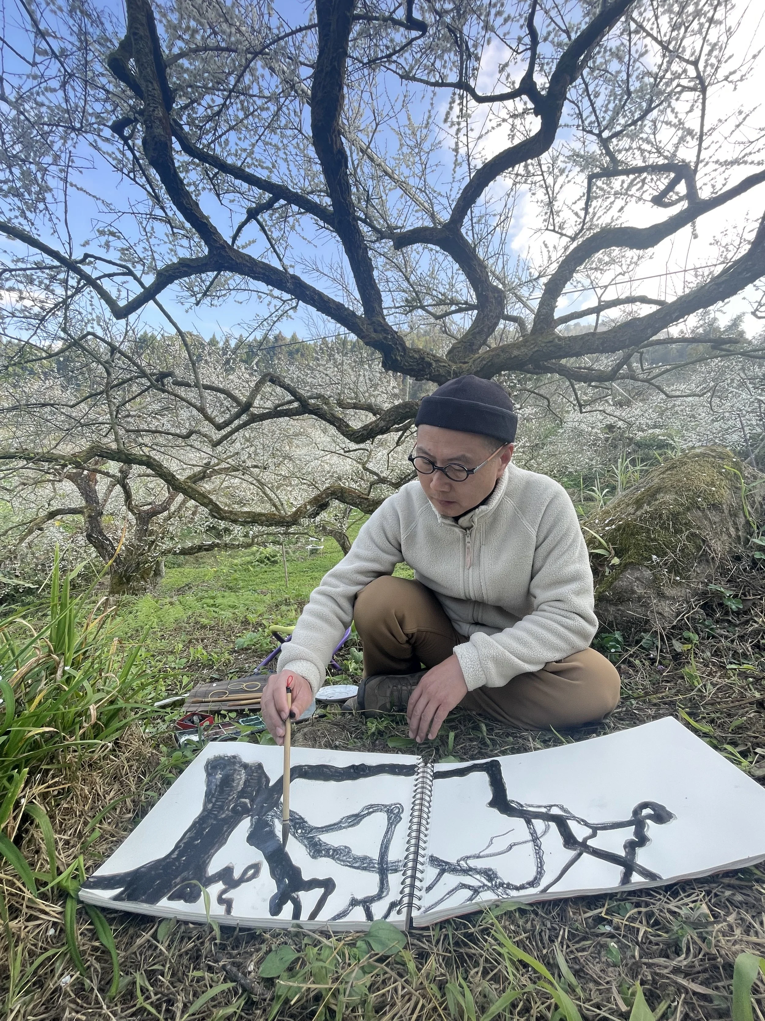 A man wearing glasses and a black beanie is sitting outdoors on the ground, surrounded by trees with spring blossoms. He is drawing on a large sketchbook with black ink, creating an abstract, branching pattern that resembles tree roots or branches. There are art supplies spread out around him.