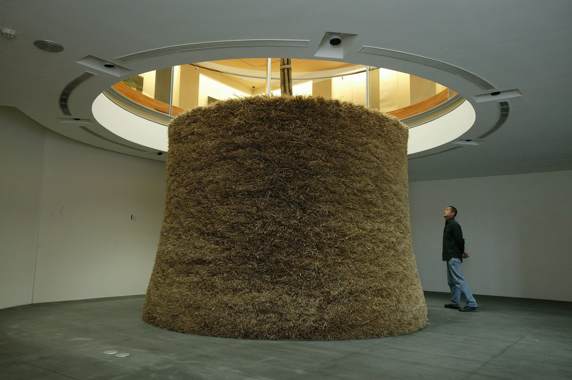 Large art installation of a tree trunk made from stacked straw or hay inside a modern gallery space. A man stands nearby looking at it.