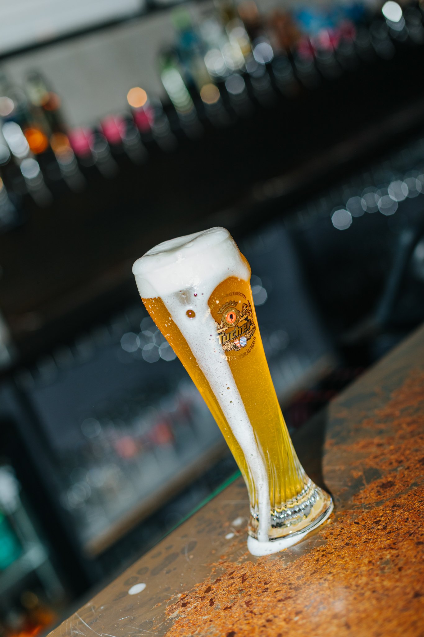 A tall glass of beer with foam on top, placed on a bar counter, with a blurred background of beer bottles.