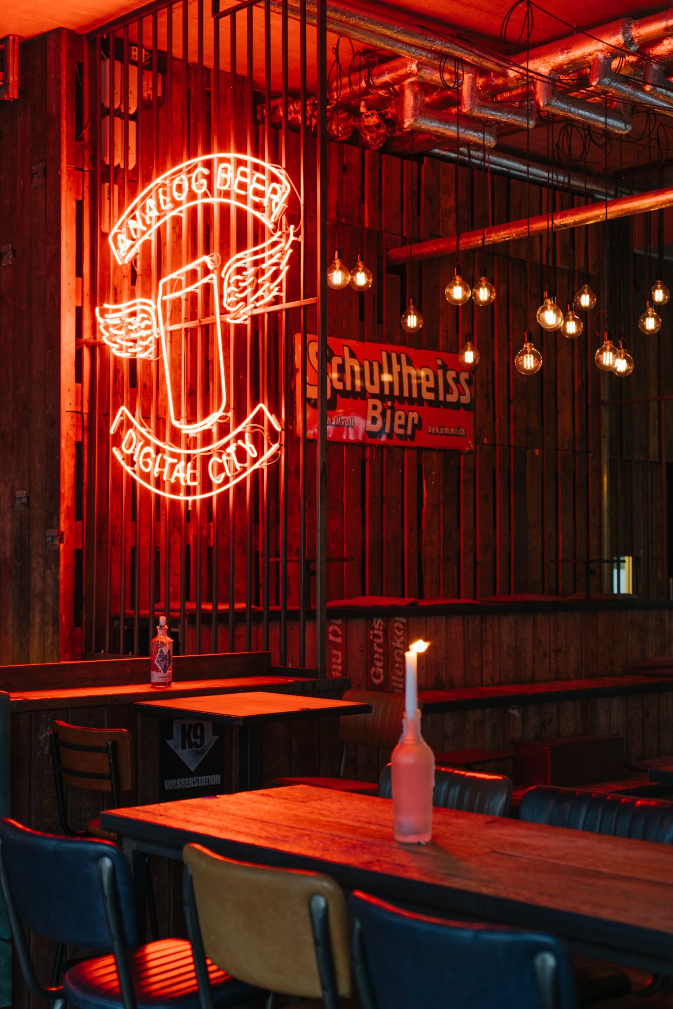 Neon sign hangs in a dimly lit bar with exposed pipes and hanging light bulbs. A table in the foreground has a candle and a bottle.