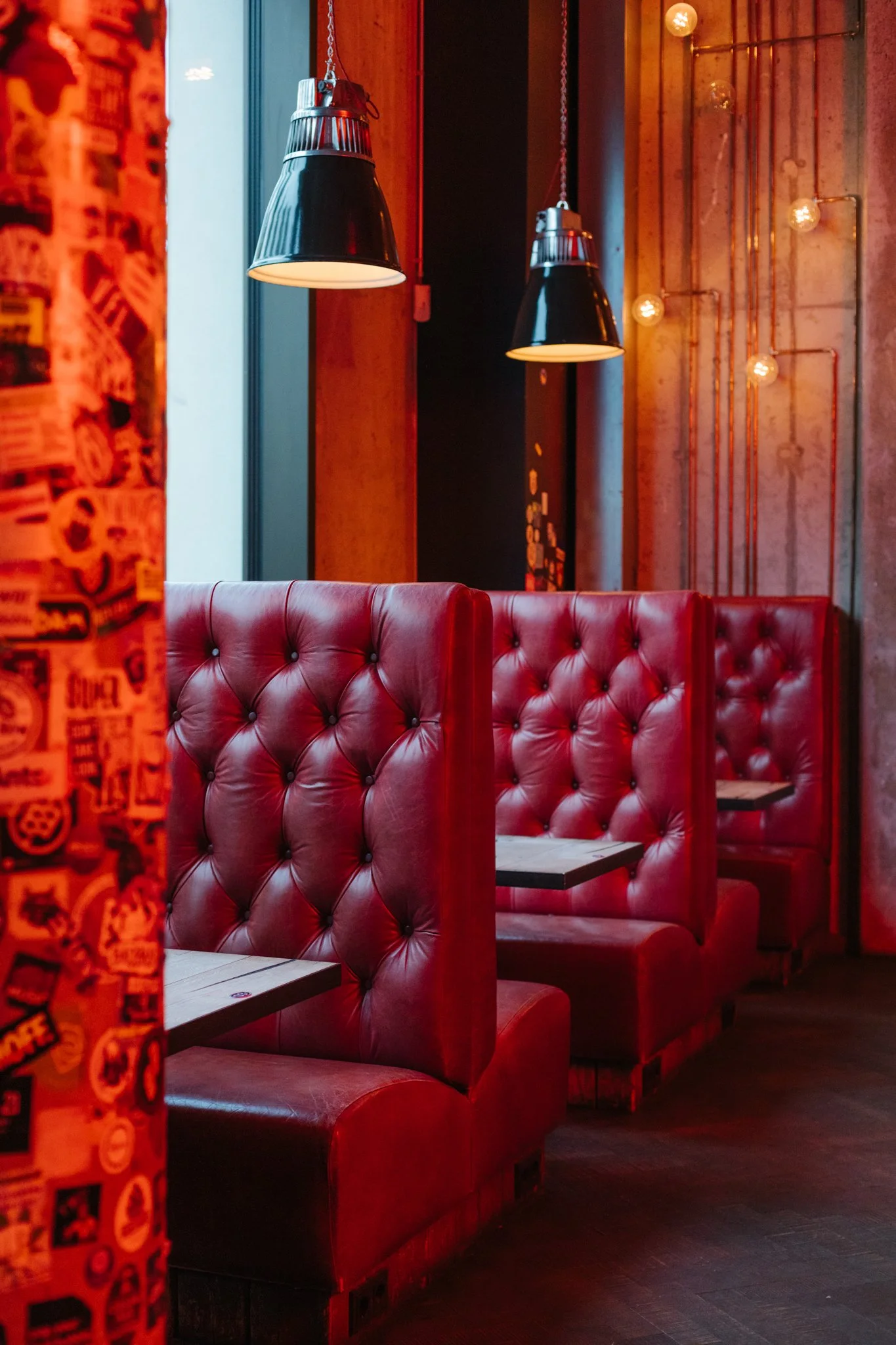 Red leather tufted booth seating with small tables in a dimly lit bar, with black hanging pendant lights and warm-colored wall lighting.