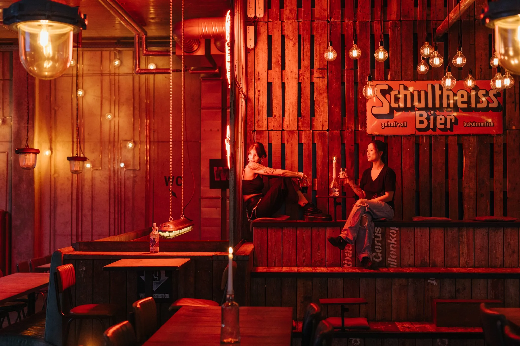 Inside a dimly lit bar decorated with warm red lighting and Edison-style hanging light bulbs. Two women are seated, one with tattoos on her arm, the other with long hair, engaging in conversation. The background features a wooden wall with a sign tha