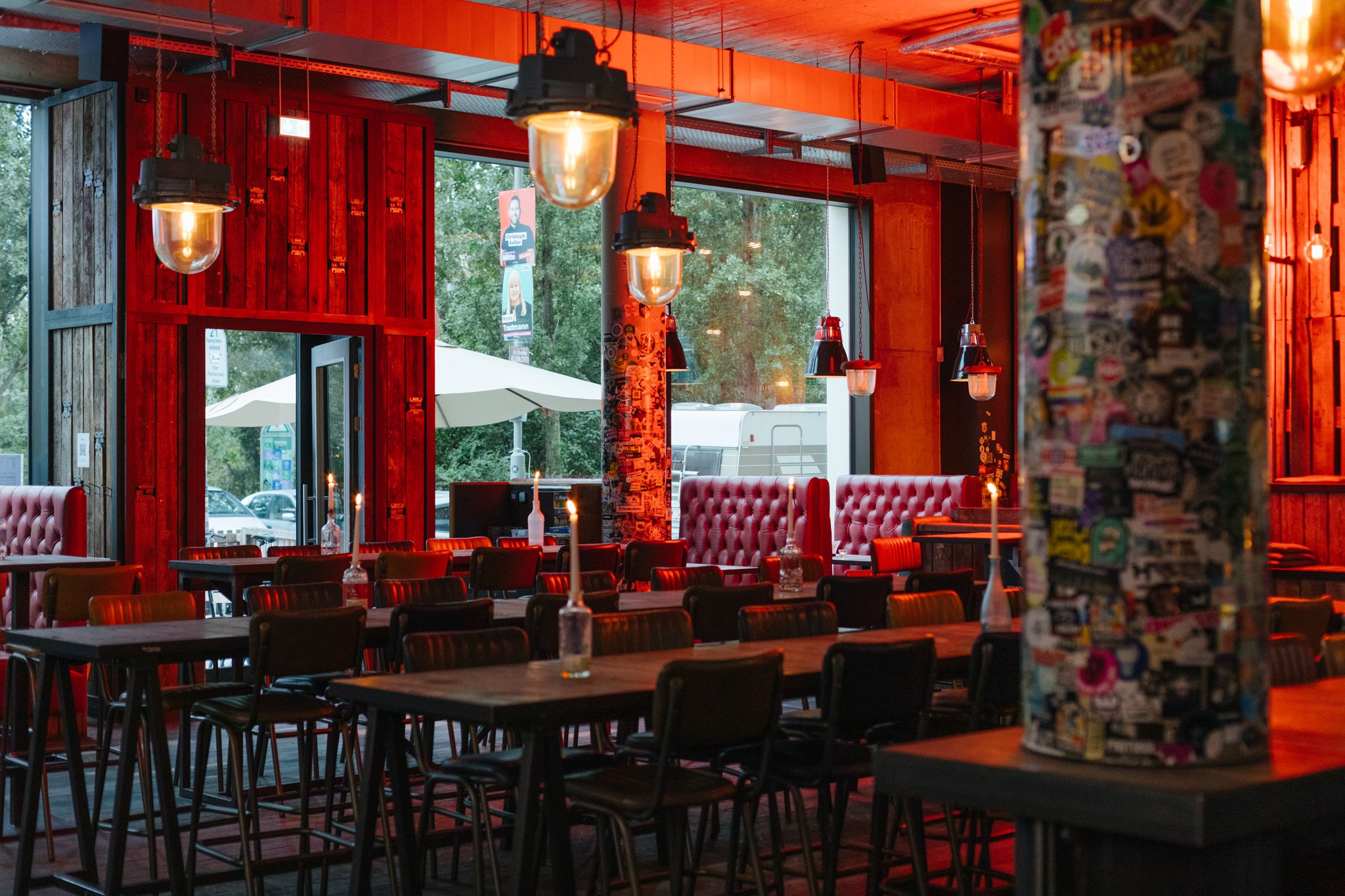 Interior of a restaurant with wooden and concrete walls, hanging pendant lights, red cushioned booths, and leather chairs around tables set with candles. Large windows show trees and umbrellas outside.