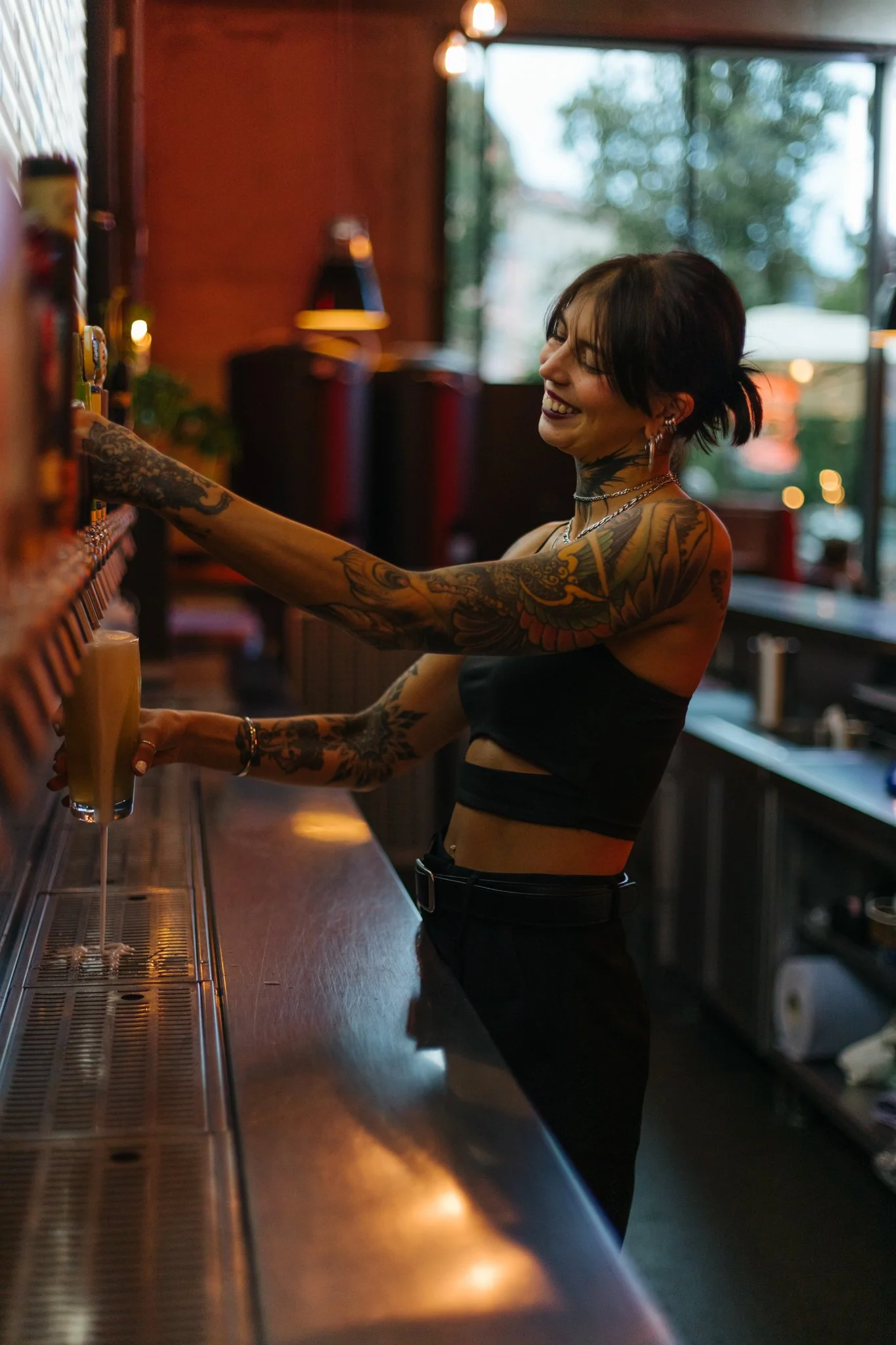 A smiling woman with tattoos on her arms and neck, wearing a black crop top and black pants, is serving drinks at a bar. She has short dark hair and is wearing earrings and layered necklaces. The bar has a warm, dimly lit atmosphere with a large wind