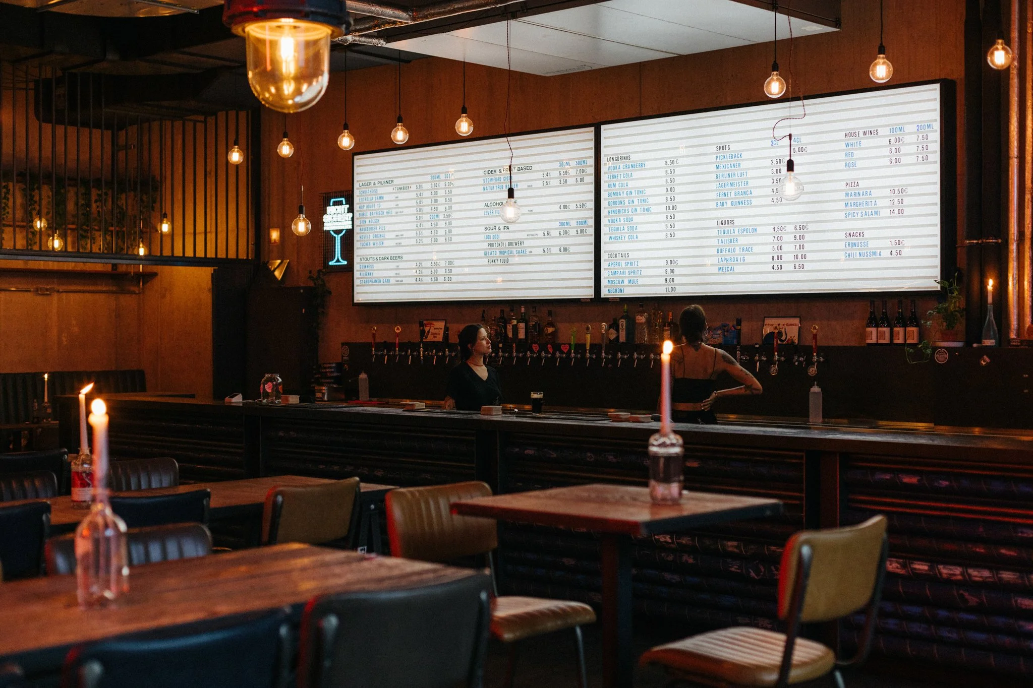 A cozy bar interior with dim lighting, candles on tables, a large digital menu board displaying drink options, and two staff members behind the bar.