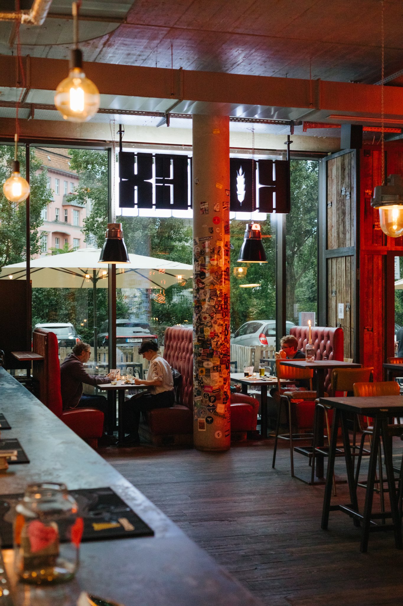 Inside of a modern restaurant with large windows, red seating, pendant lighting, patrons dining, and an outdoor patio with umbrellas visible through the windows.