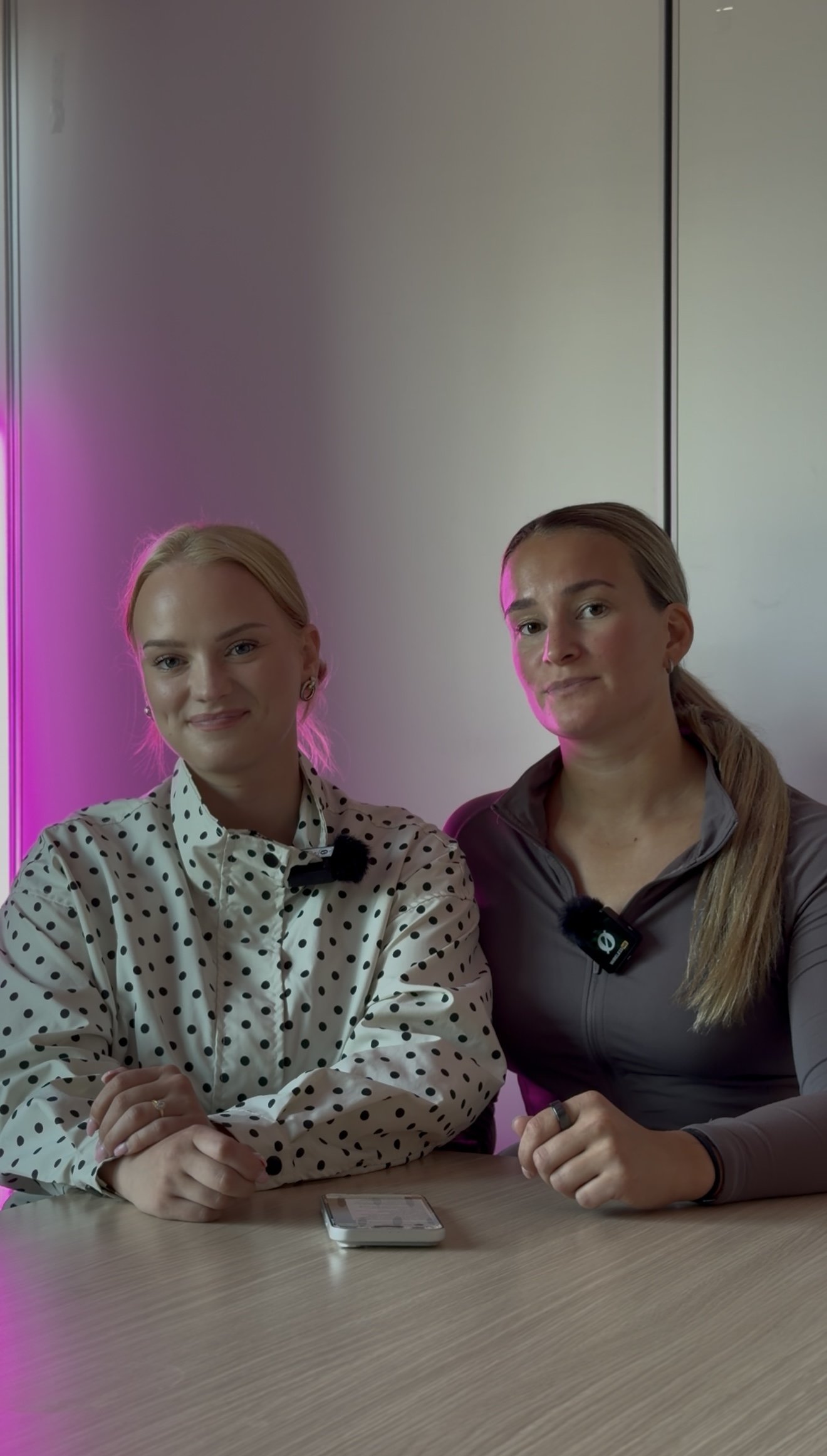 Kaksi naista istuu pöydän ääressä sisällä, taustalla harmaat seinät ja loisteputkilamppu. Molemmilla on mikrofonit kiinnitettynä yläruumiin alueelle.