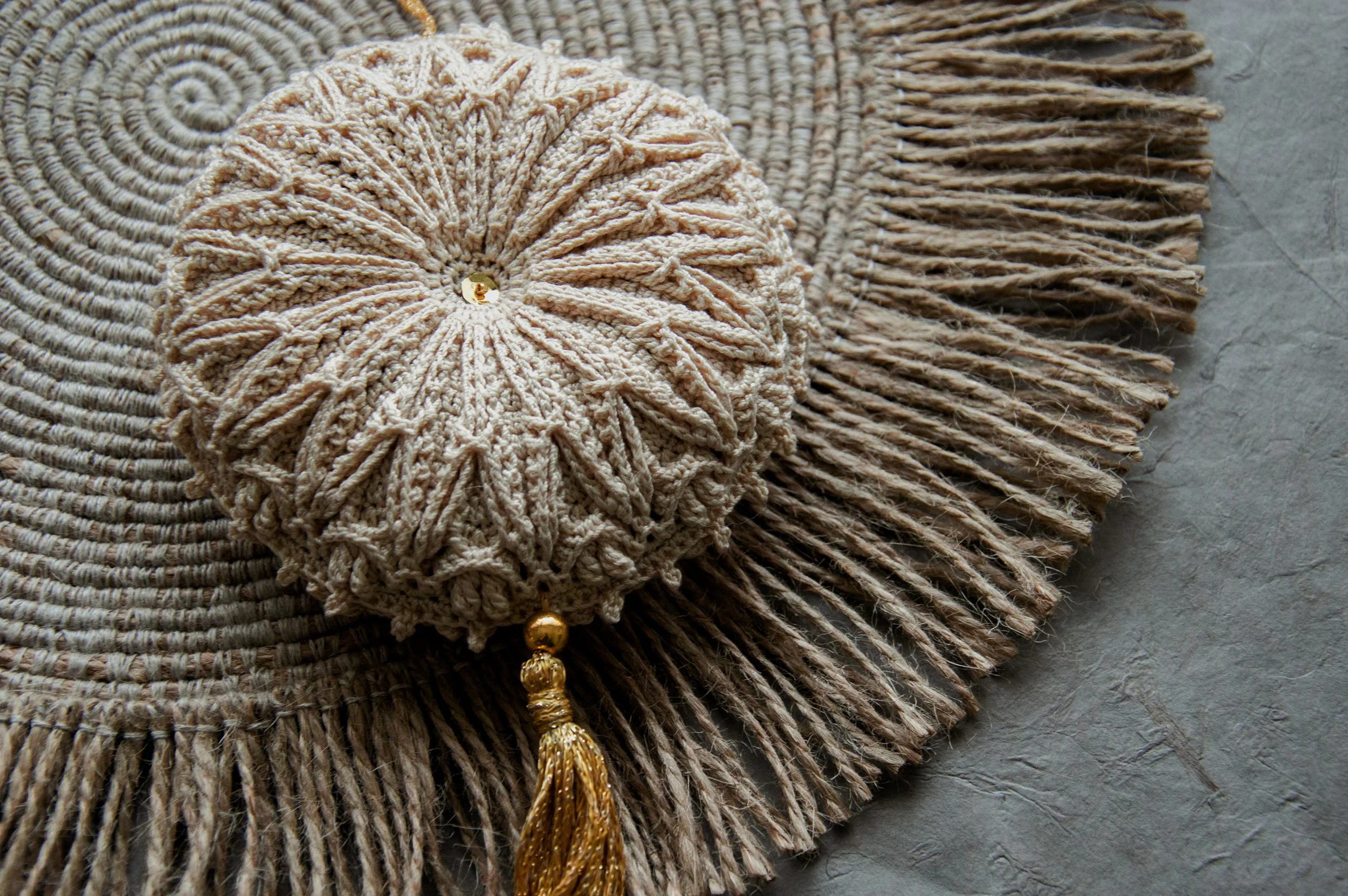 Marita crochet ornament in natural beige with a gold tassel, displayed on a woven jute mat