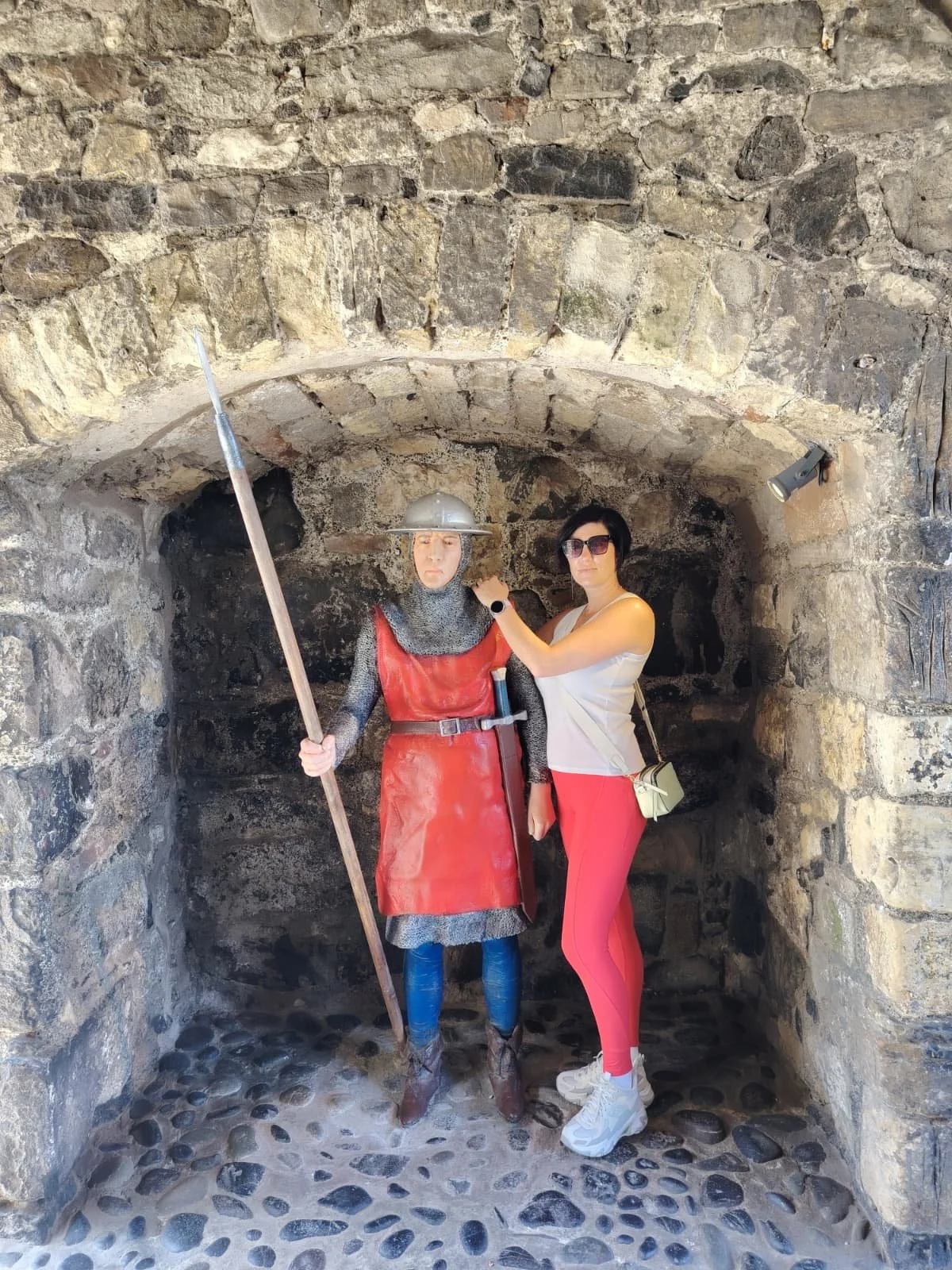 A woman with black hair, sunglasses, a white top, and red pants, standing next to a medieval knight statue made of metal, in a stone archway.